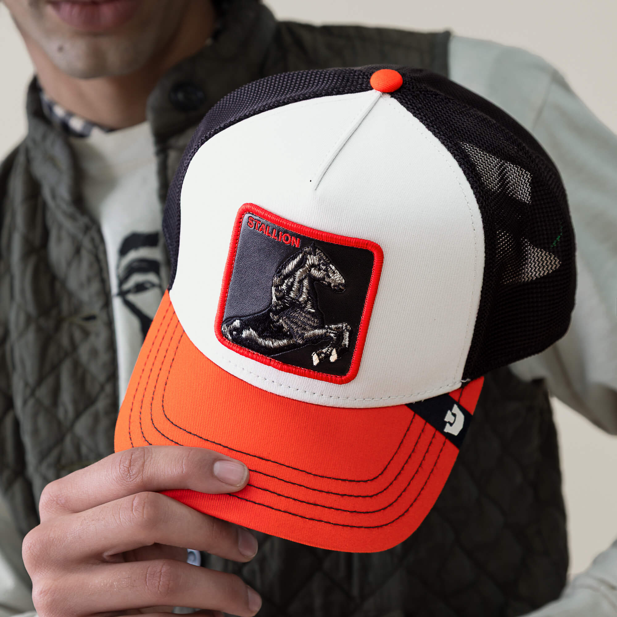 Someone holds the Goorin Bros. Tricolor Stallion trucker hat, which has a white front, black mesh back, bright orange visor, and an embroidered stallion patch labeled STALLION.