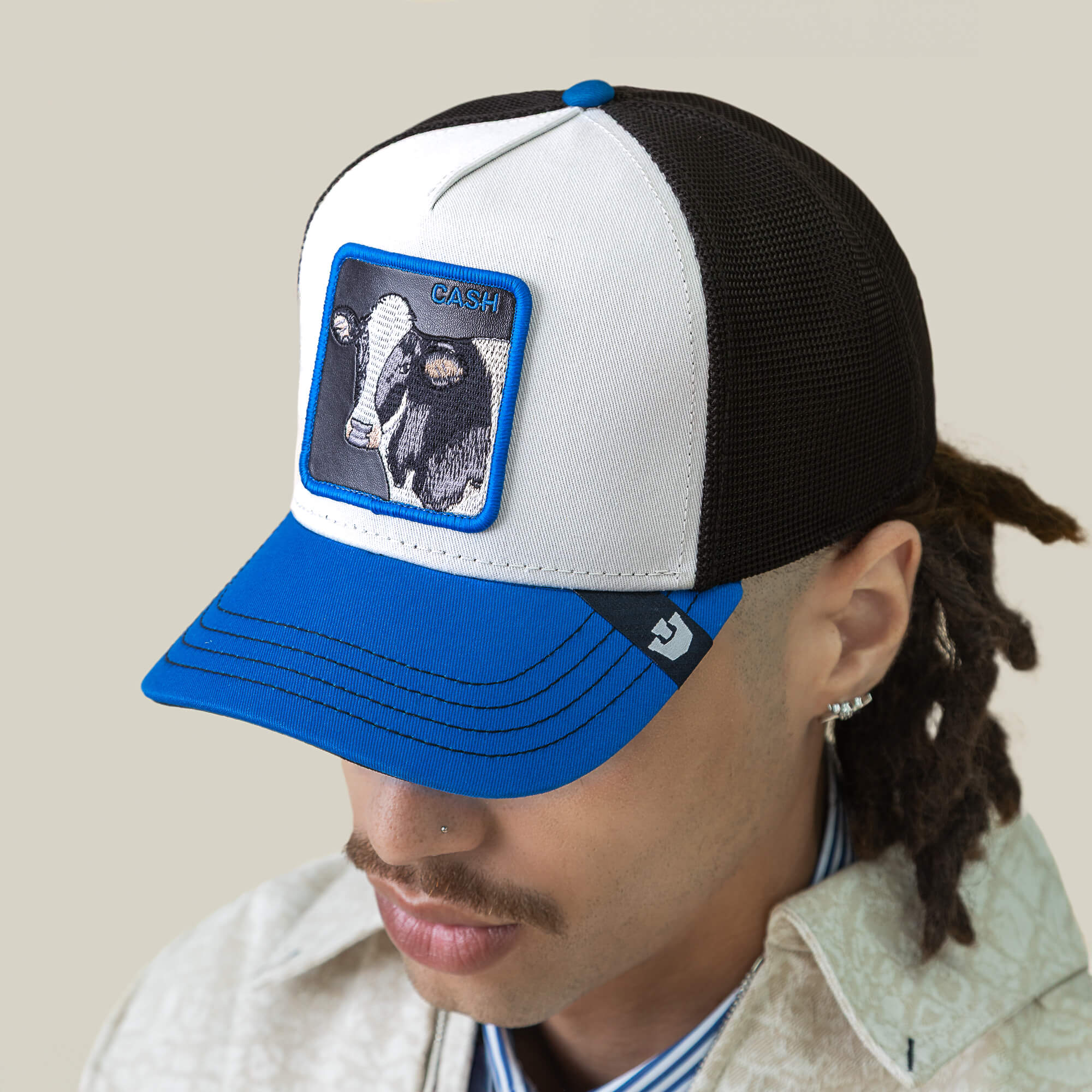 A person wears the Goorin Bros. "Tricolor Cow" mesh cap with a cow and CASH patch, looking downward and accessorized with facial hair, earrings, and a stylish wallet.