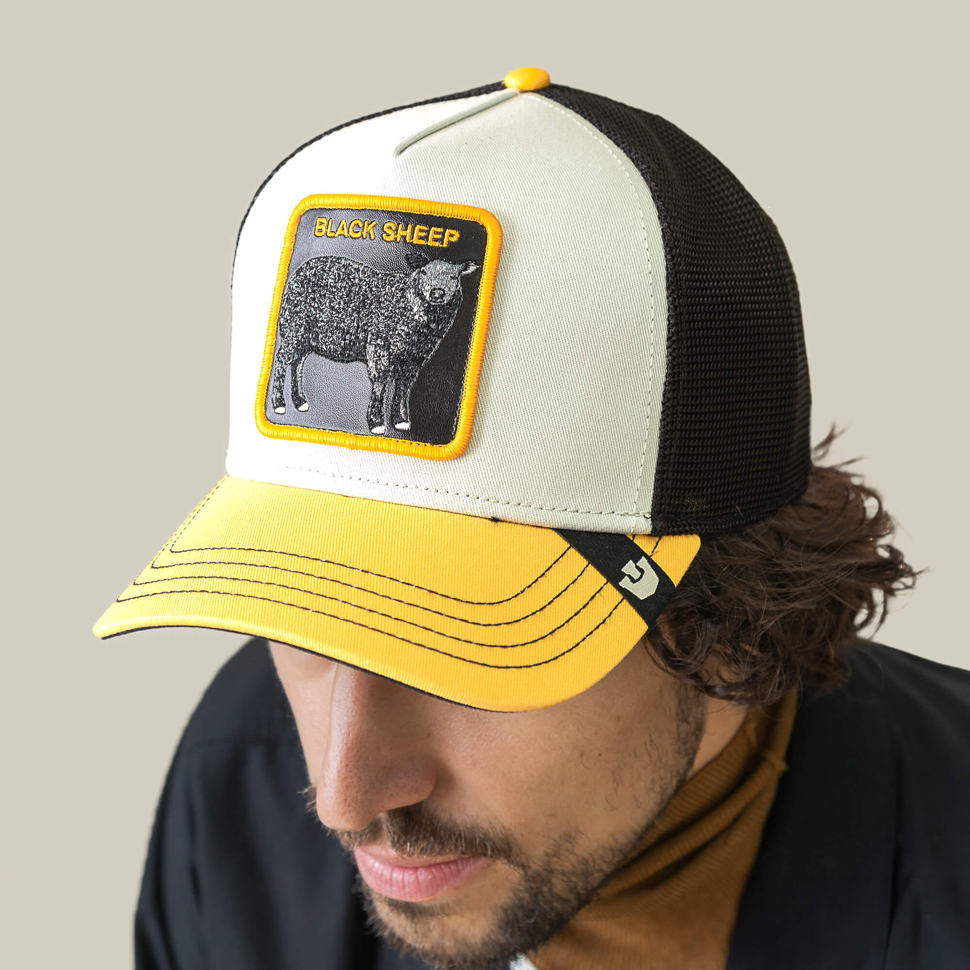 A person wears the Goorin Bros. Tricolor Sheep hat, a bold black mesh cap featuring a white, yellow, and black design with a statement patch displaying BLACK SHEEP and a black sheep illustration.