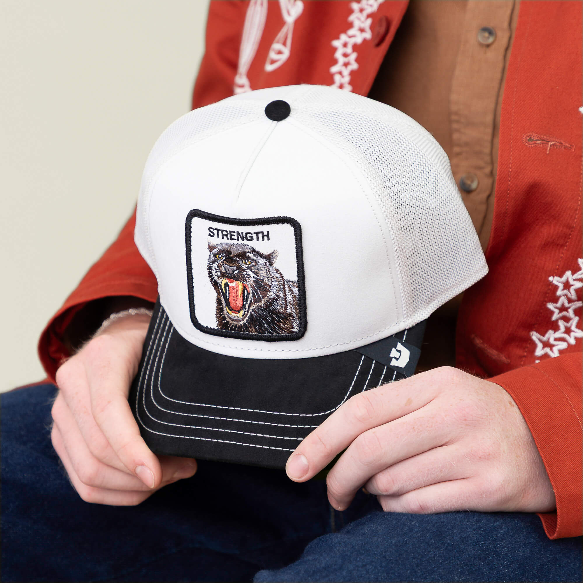 A person in a red jacket holds the Goorin Bros. “Suede Panther” cap, featuring a white crown, black brim, and a panther patch labeled "STRENGTH.