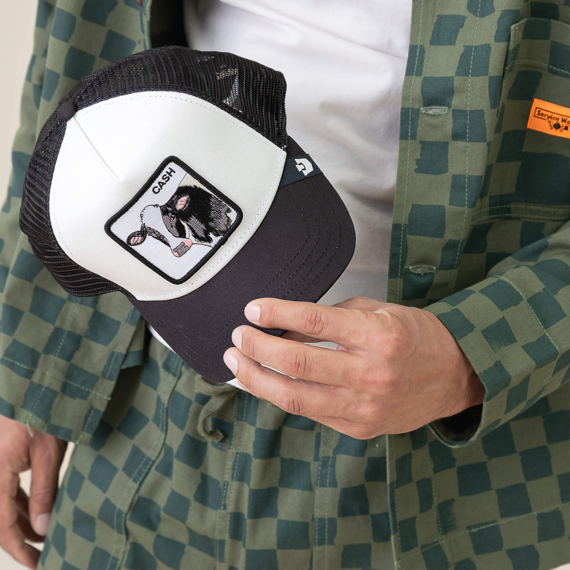 A person in a green checkered jacket holds the Goorin Bros. Cash Cow Trucker hat, featuring a black and white design with a bold bovine cash register patch labeled CASH—capturing the spirit of the Cash Cow.