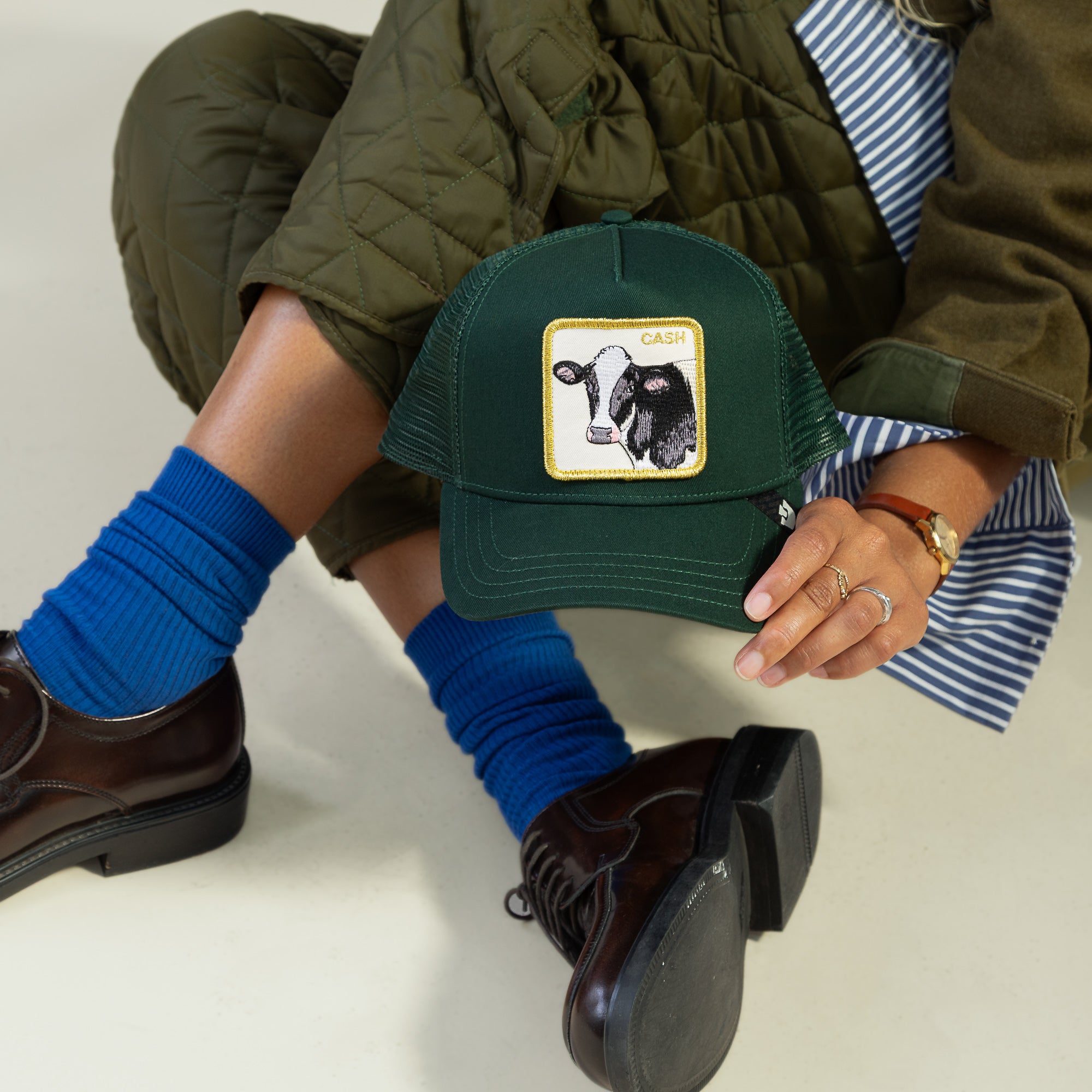 A person sits cross-legged in blue socks, brown shoes, and a green quilted coat, holding the Goorin Bros. Cash Cow Trucker cap—green with a bold bovine patch and the word "CASH.