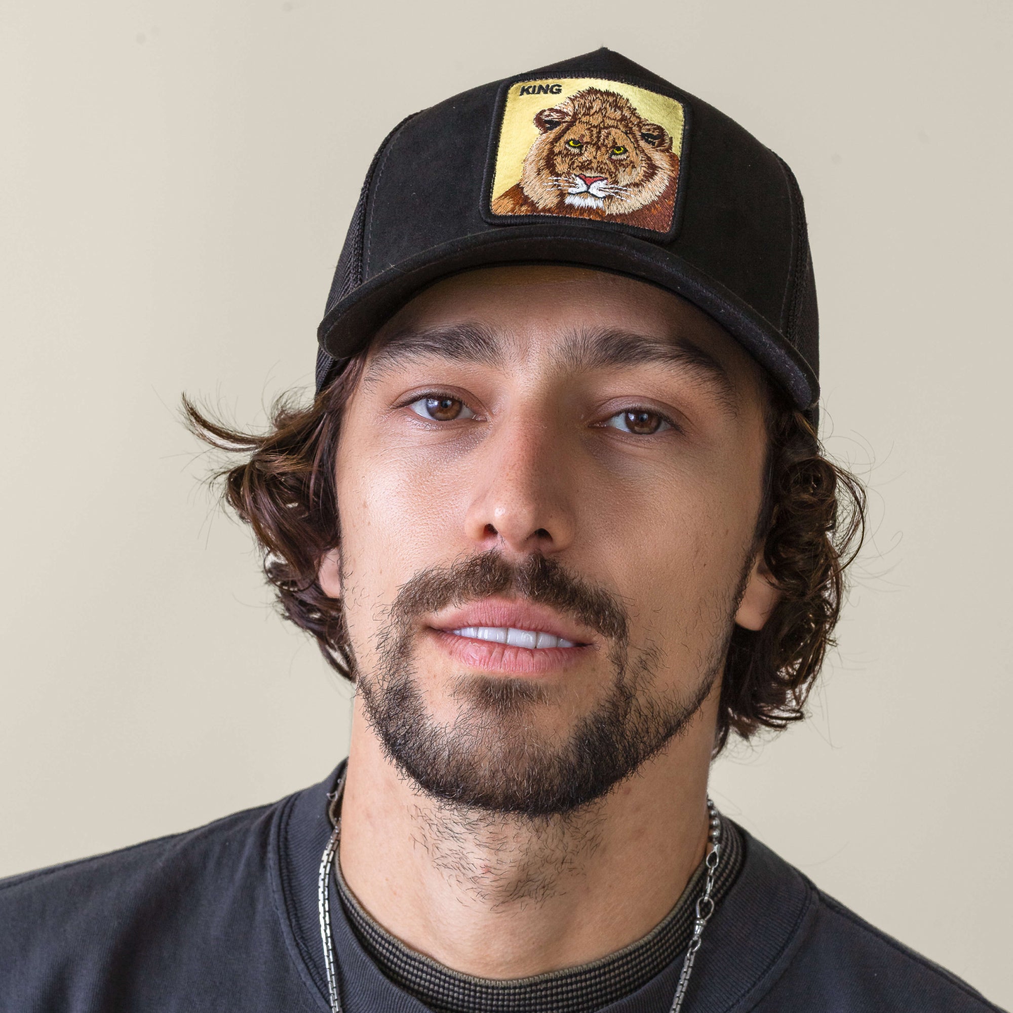 A man with a mustache and beard wears the Goorin Bros. Golden King black cap with a Golden Lion patch and "KING" text, paired with a black shirt and silver necklace, against a plain background.