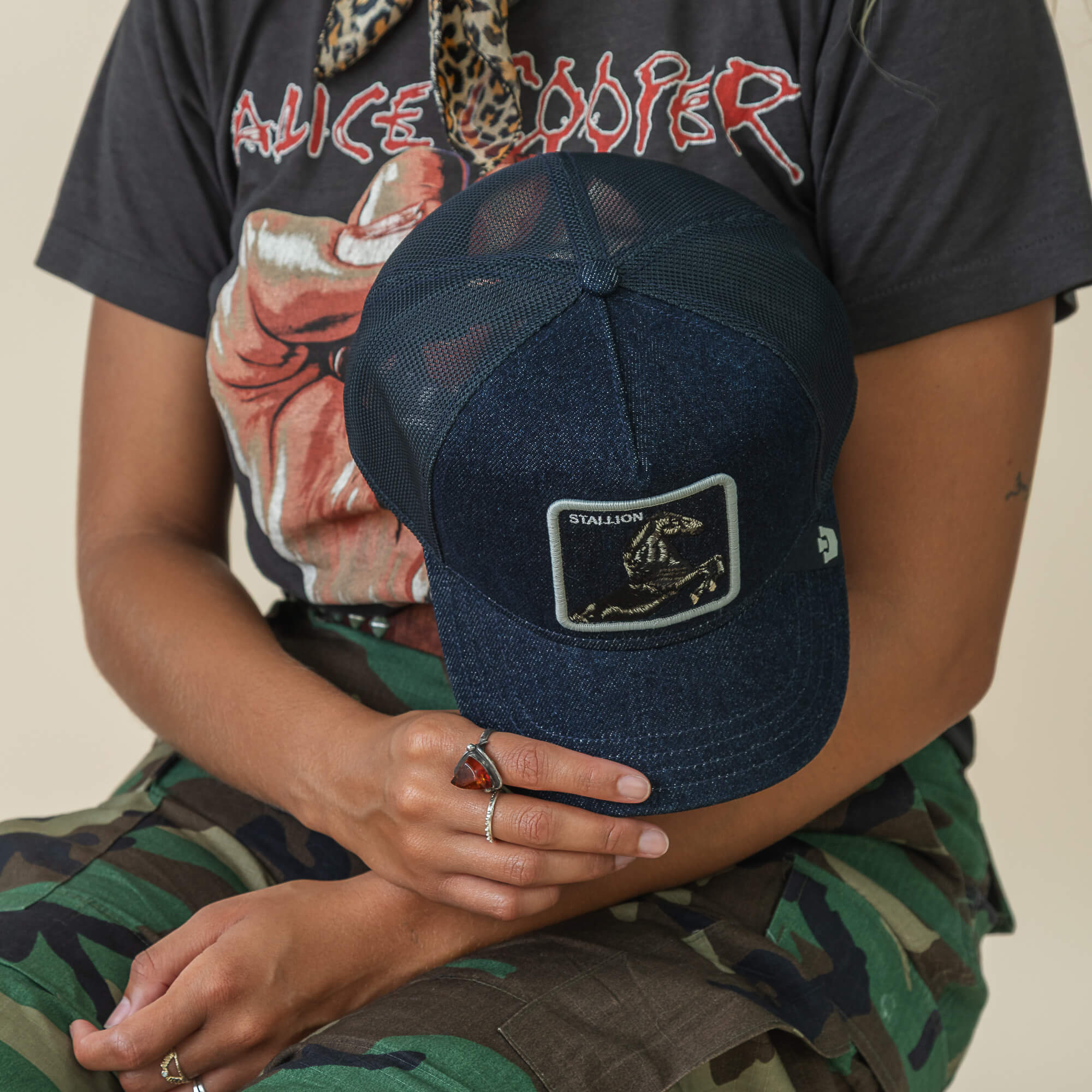 A person in a black Alice Cooper t-shirt and camouflage pants holds a Goorin Bros. Denim Stallion blue embroidered cap with a rectangular patch in their right hand.