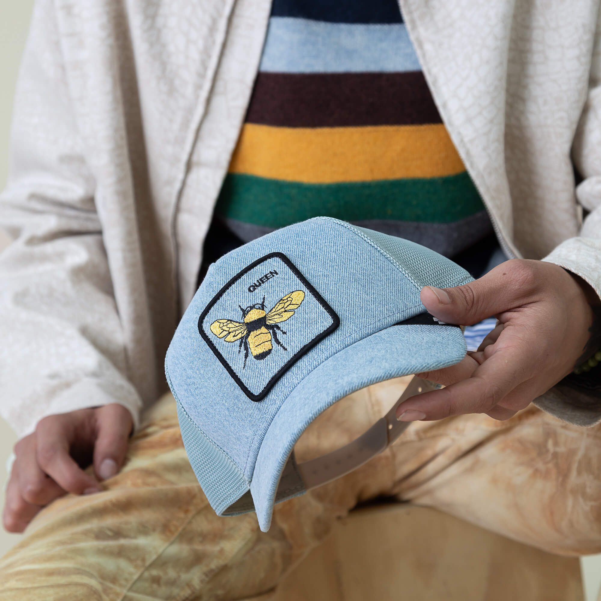 A person holds a light blue Denim Queen cap by Goorin Bros., featuring a yellow bee patch and the word QUEEN, while wearing patterned pants and a striped sweater under a light jacket.