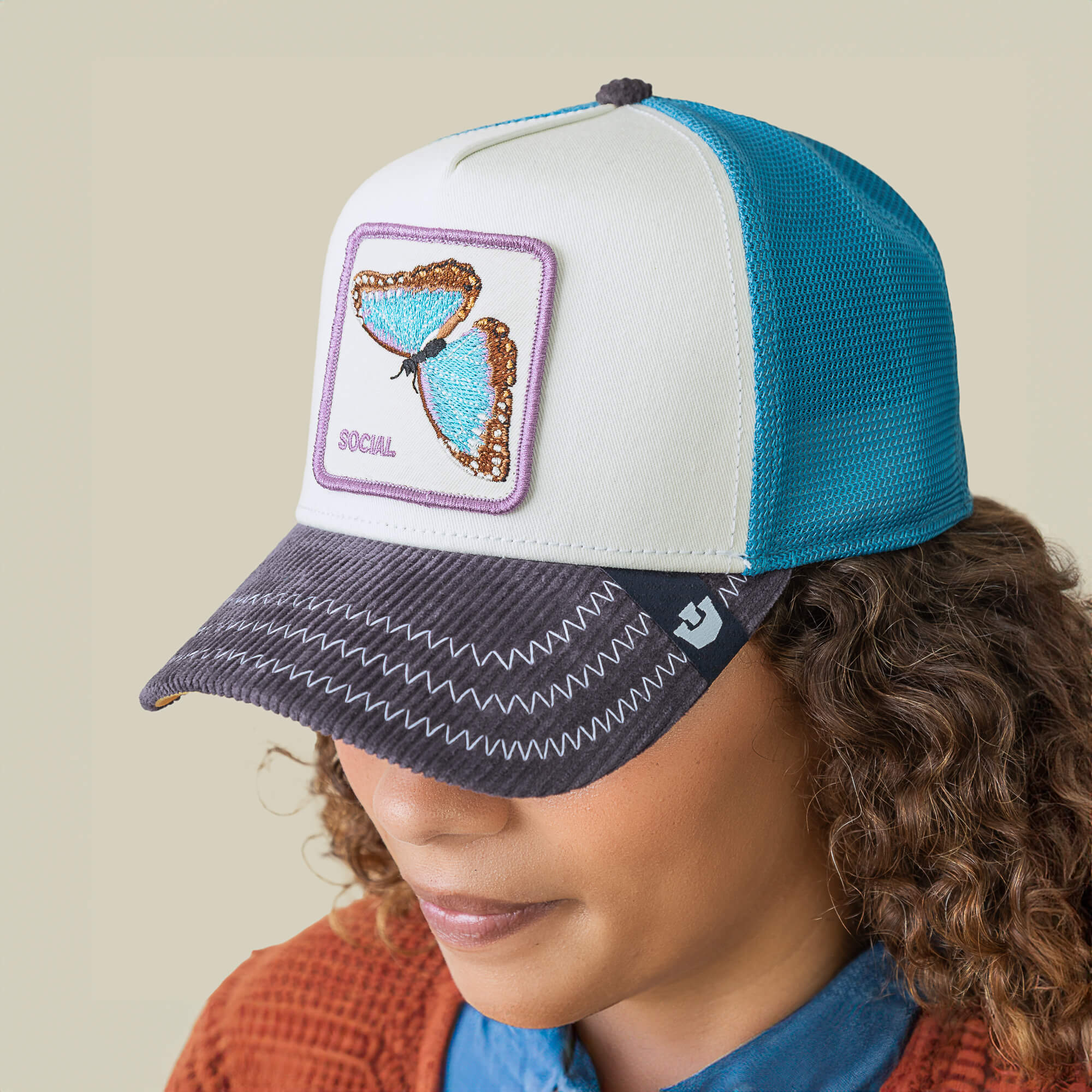 A person wears the Goorin Bros. Social Remix hat, featuring blue and white panels, a butterfly patch, “SOCIAL” on the front, a corduroy brim, and visible zigzag stitching for a unique look.