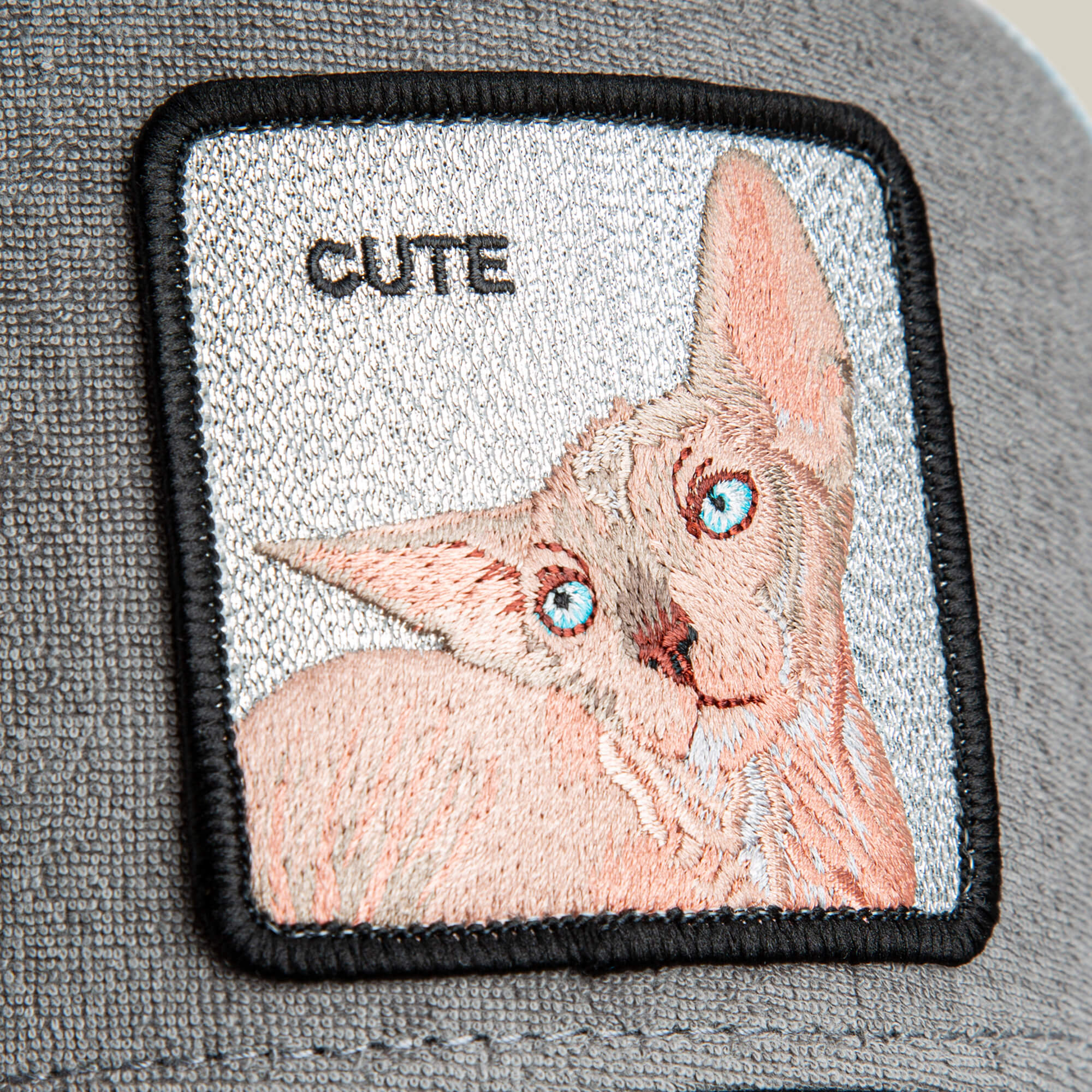 A close-up of the Goorin Bros. "Cute Remix" patch, featuring a gray terry cloth background, an embroidered blue-eyed hairless cat, and "CUTE" stitched above it.