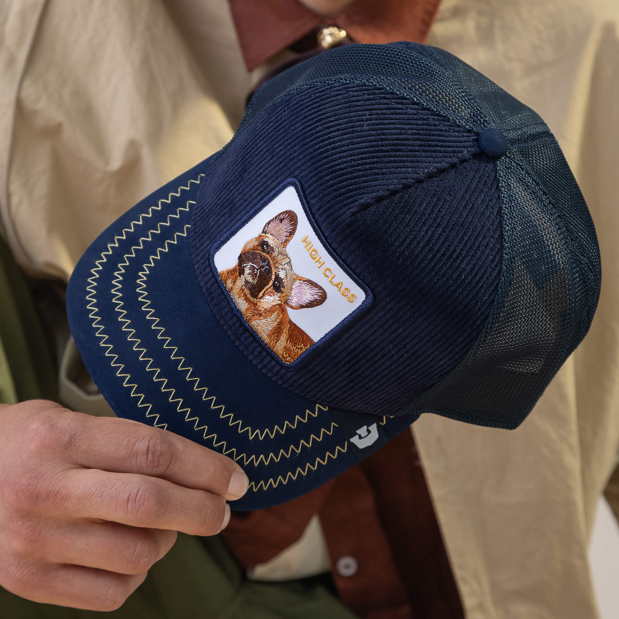 A person holds the Goorin Bros. High Class Remix cap, a navy blue mesh hat with an embroidered dog patch and "HIGH CLASS" text, its navy textures adding depth.