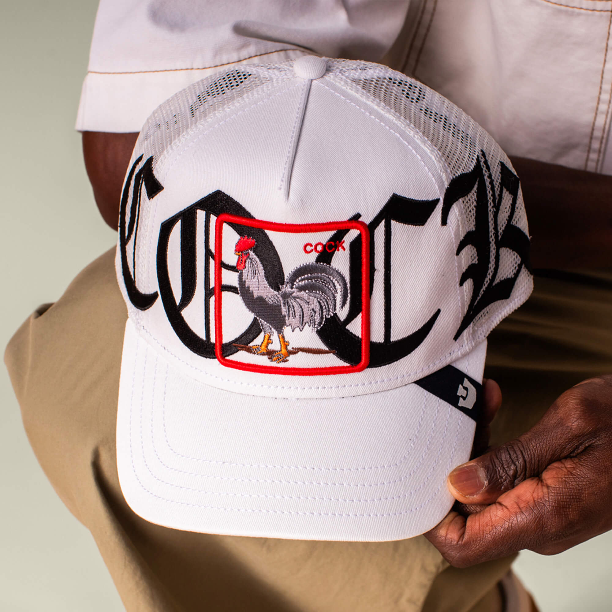 A person holds the Goorin Bros. Old English Cock cap, a white mesh baseball hat featuring a rooster patch labeled COCK and bold Old English script in black and white across the front.