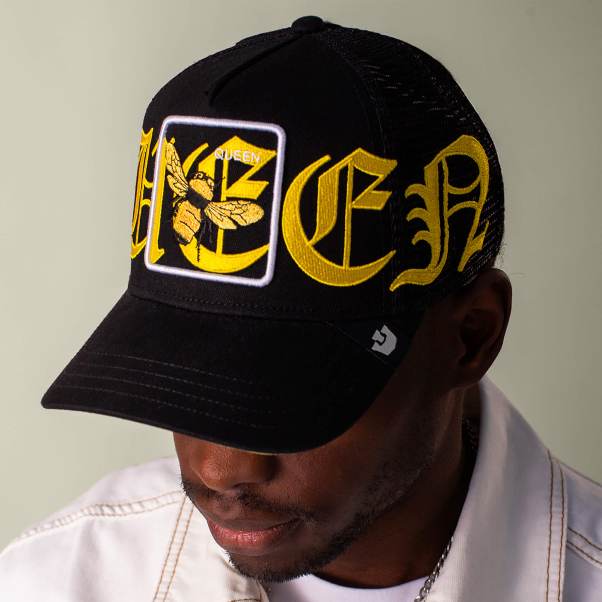 A person sports the Goorin Bros. Old English Queen black cap, featuring QUEEN in bold lettering and a queen bee patch, paired with a white jacket for a standout black and yellow look.