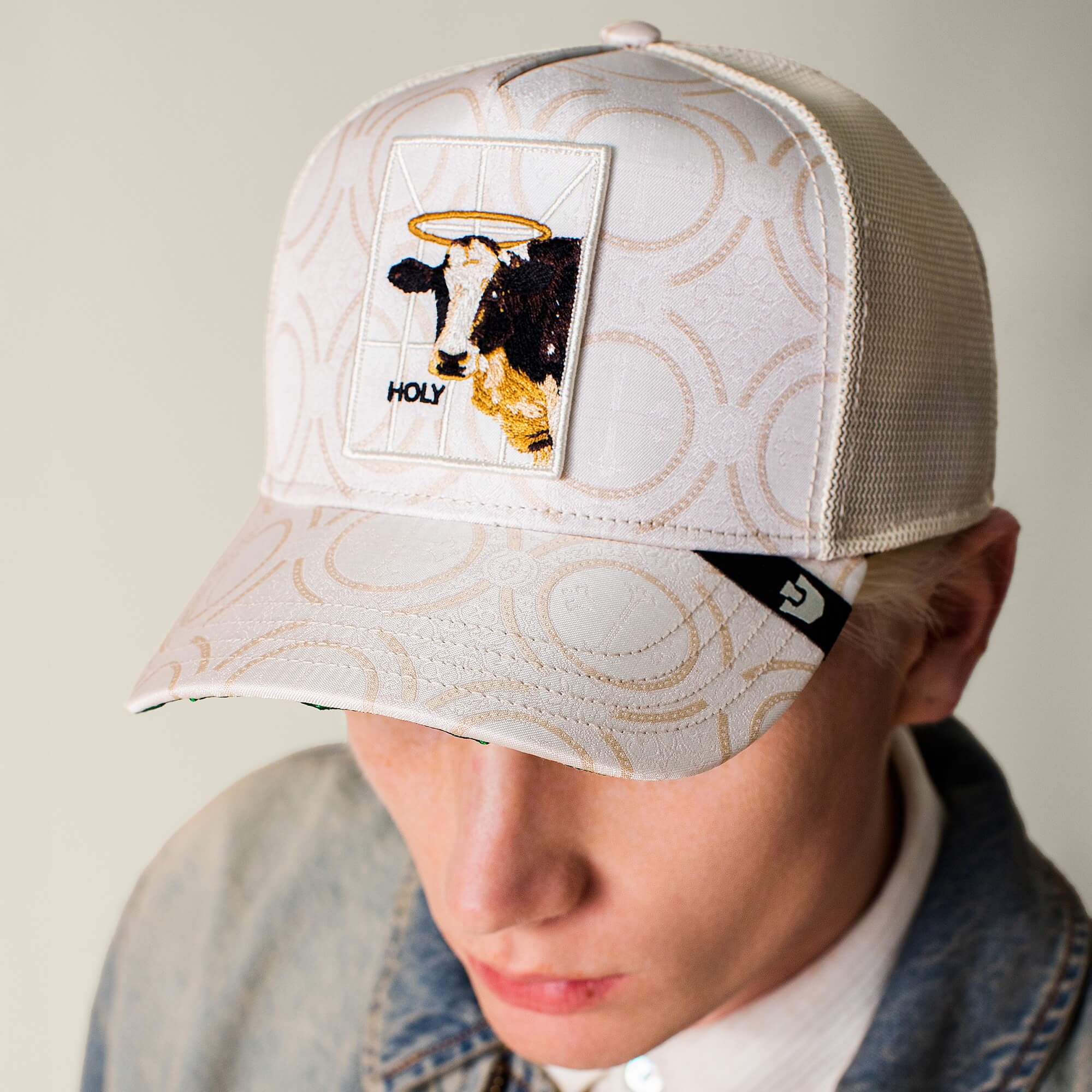 Someone wears the Goorin Bros. "Religulous - Holy Cow" light baseball cap, featuring a patch of a sacred cow with a halo and the word “HOLY”—bringing together faith and humor. The background is plain and out of focus.