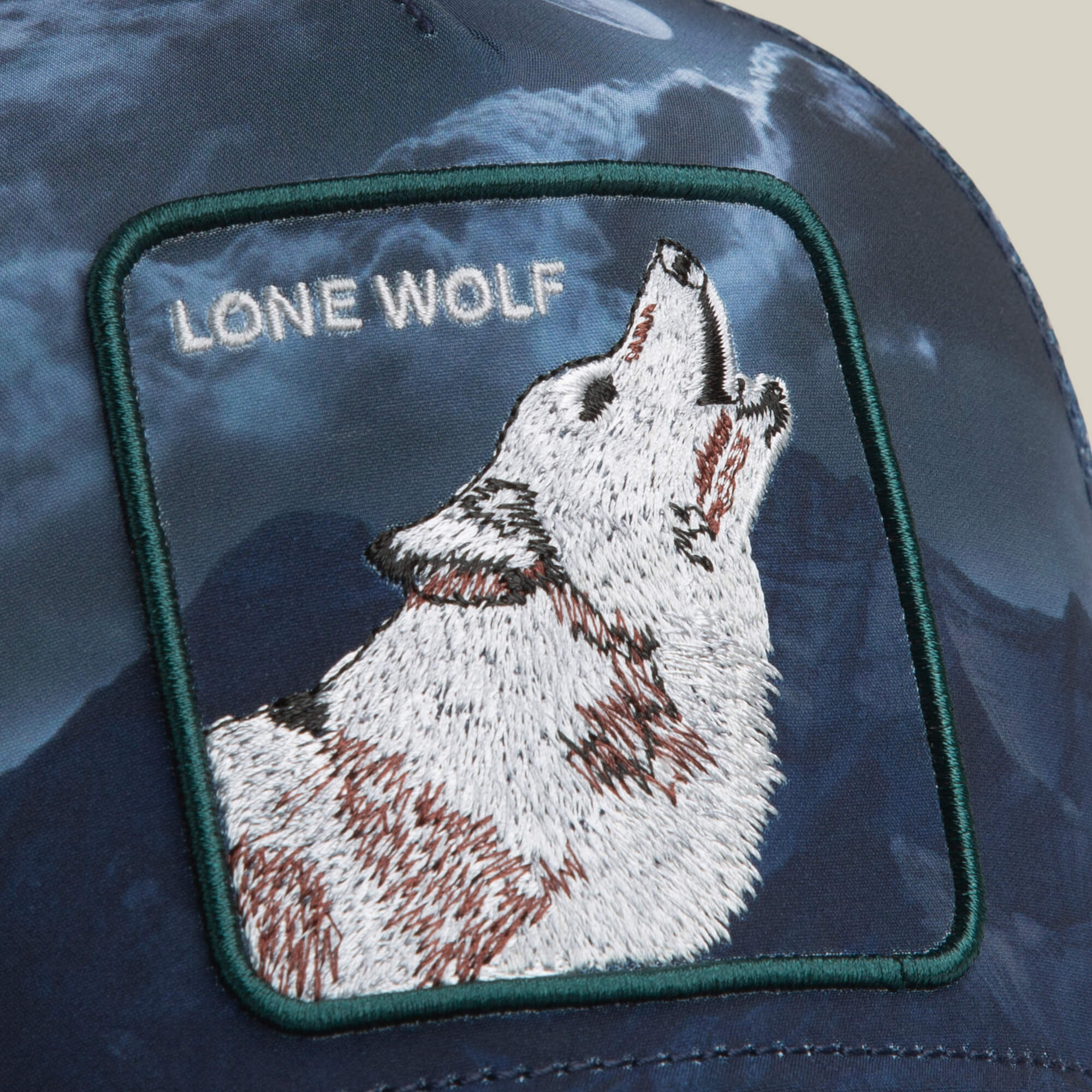 A close-up of a Goorin Bros. “Wolf In The Element” trucker hat with an embroidered howling wolf patch and text, set against a dark, mountainous background.