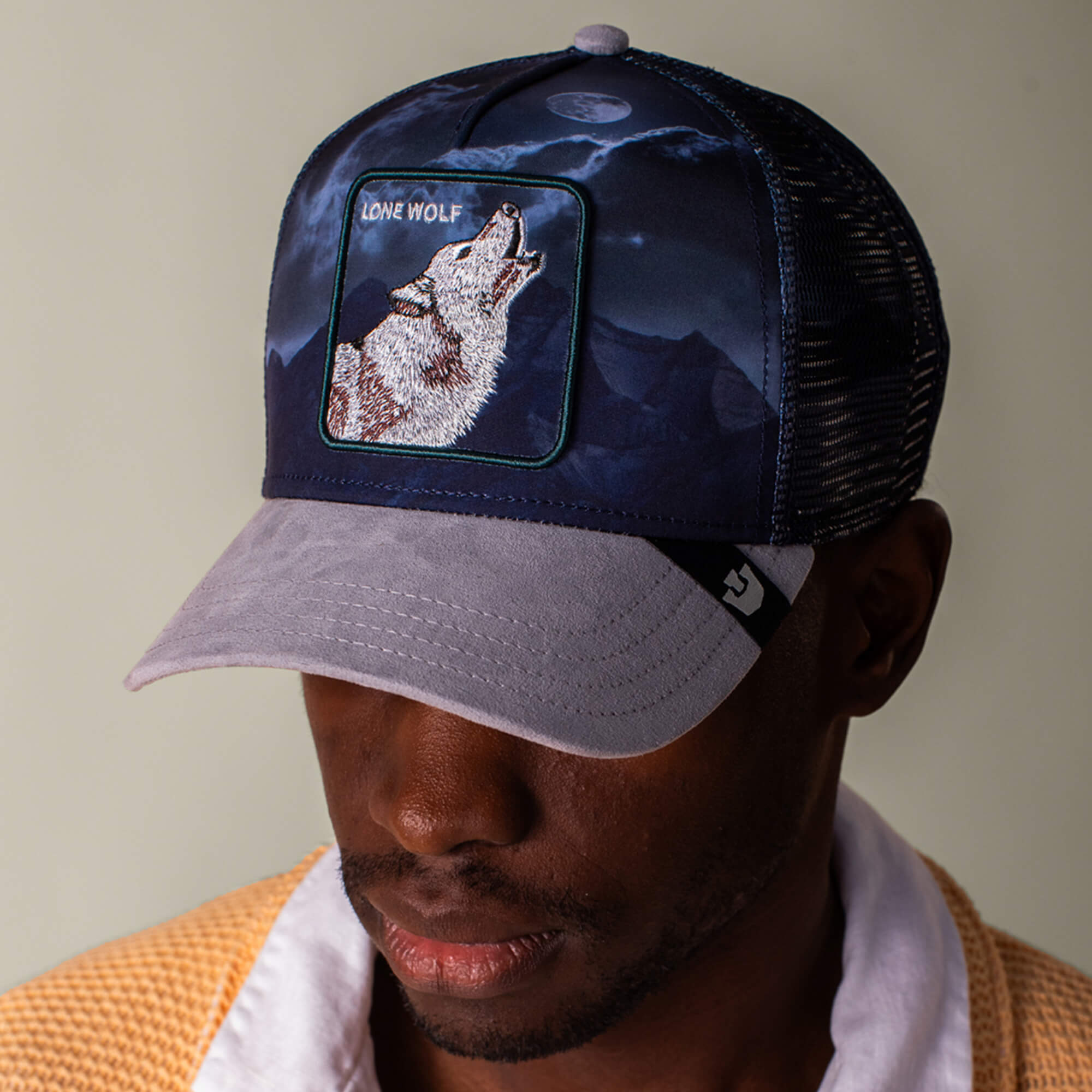 A man wearing the Goorin Bros. "Wolf In The Element" trucker hat, featuring a howling wolf patch and mountain design in navy and grey.