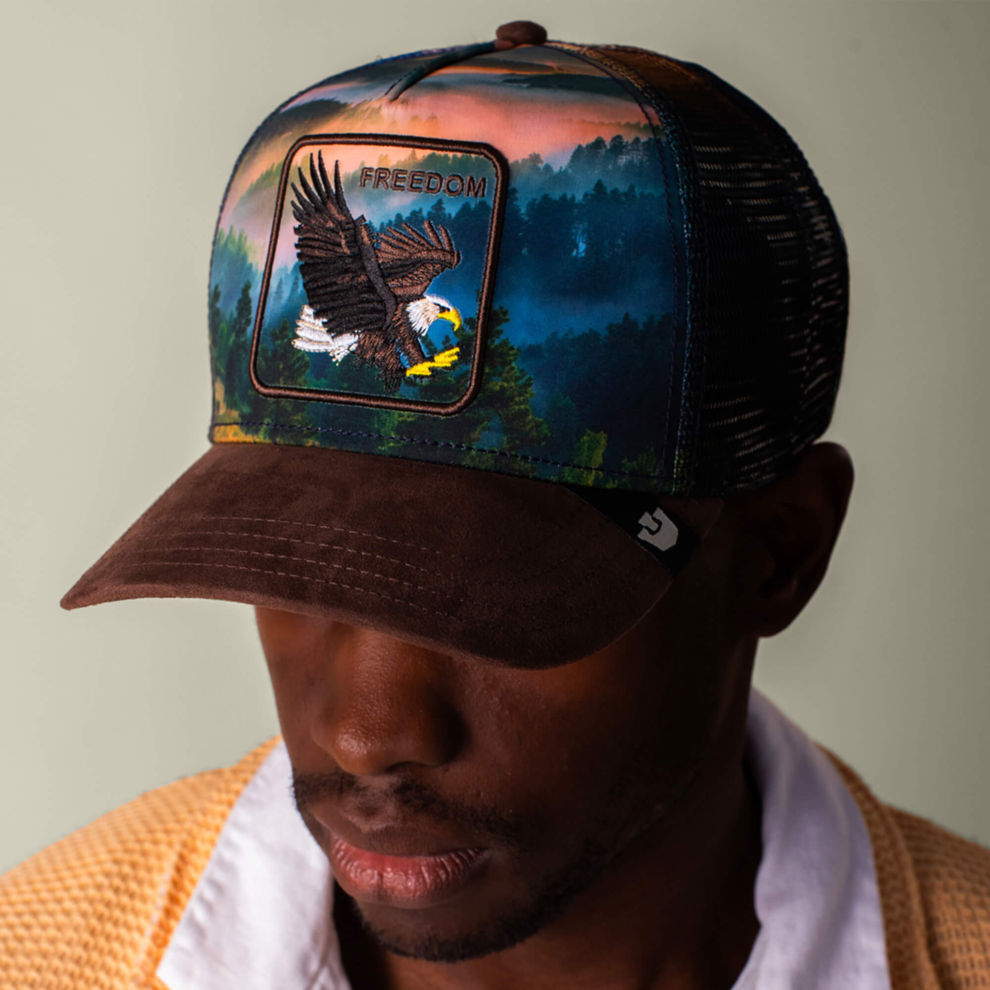 A man wears Goorin Bros.' "Eagle In The Element" cap, featuring an earth-toned landscape design, a brown brim, and a patch with an eagle and FREEDOM embroidered on it.