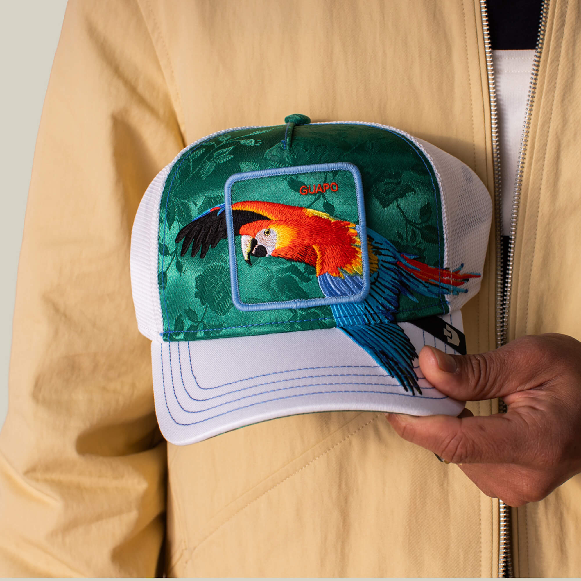 A person holds a green and white Goorin Bros. Honduras Macaw trucker hat with an embroidered tropical macaw patch and "GUAPO," while wearing a beige jacket.
