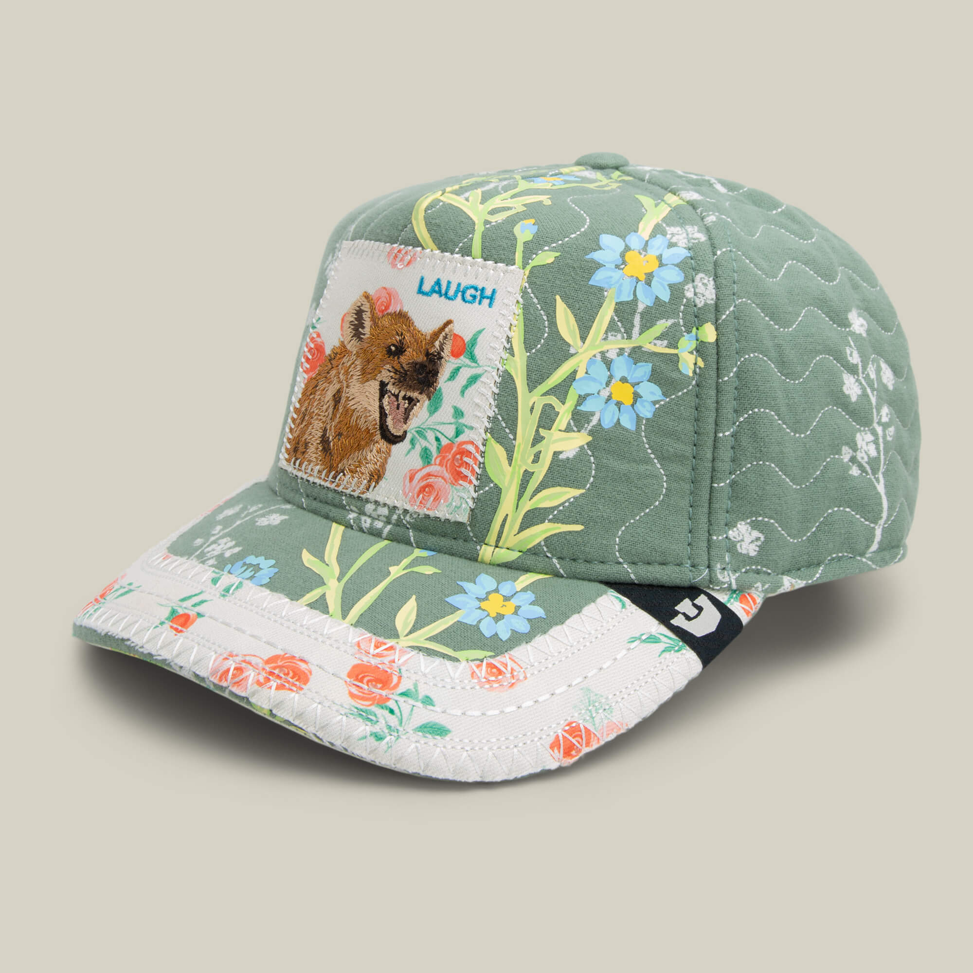 The Goorin Bros. Vol. 1 - Quiltbag: Laugh is a green floral trucker hat with patchwork patterns and a front patch of a laughing hyena and the word "LAUGH.
