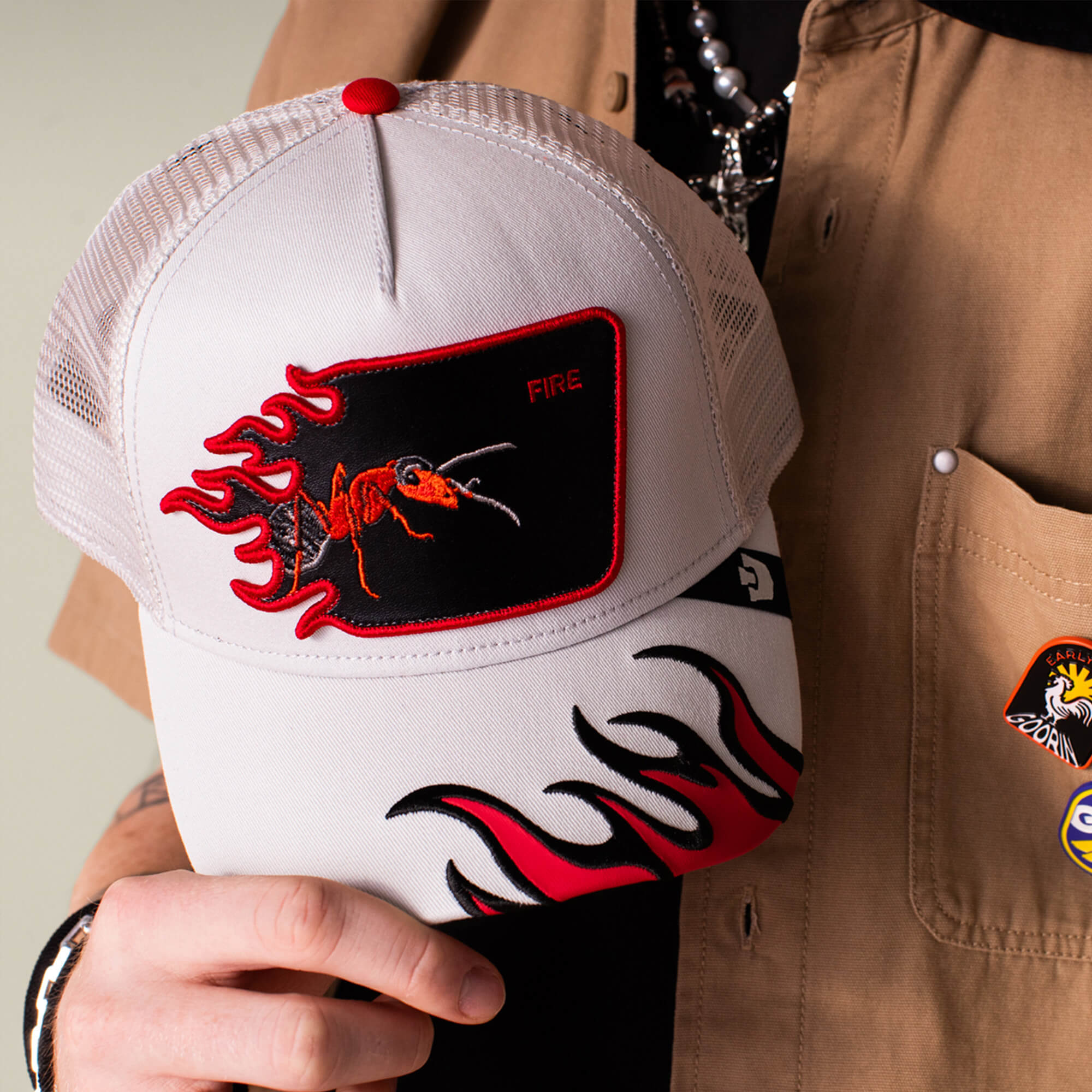 A person holds the Goorin Bros. Fire Flames trucker hat, featuring a white mesh design, red flame graphics, and a red ant patch labeled "FIRE." They are wearing a tan shirt decorated with patches.