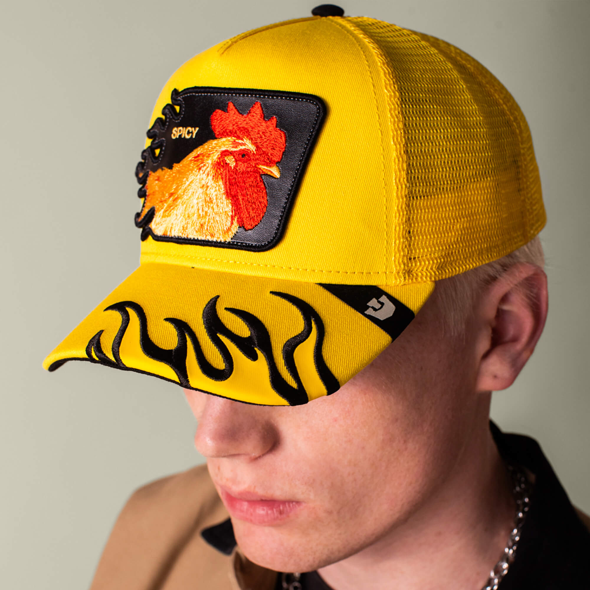A person wears the Goorin Bros. Spicy Flames trucker hat, featuring a yellow design with black flames on the brim and an embroidered rooster patch labeled "SPICY." The hat partially obscures their face.