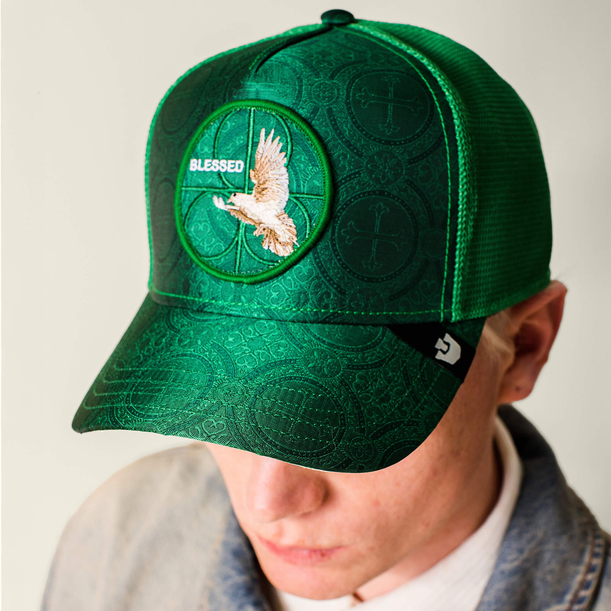 A person wears the Religulous - Blessed cap by Goorin Bros., featuring a deep green color, flying bird patch, “BLESSED” on the front, textured pattern, and a logo tag on the side for stylish detail.