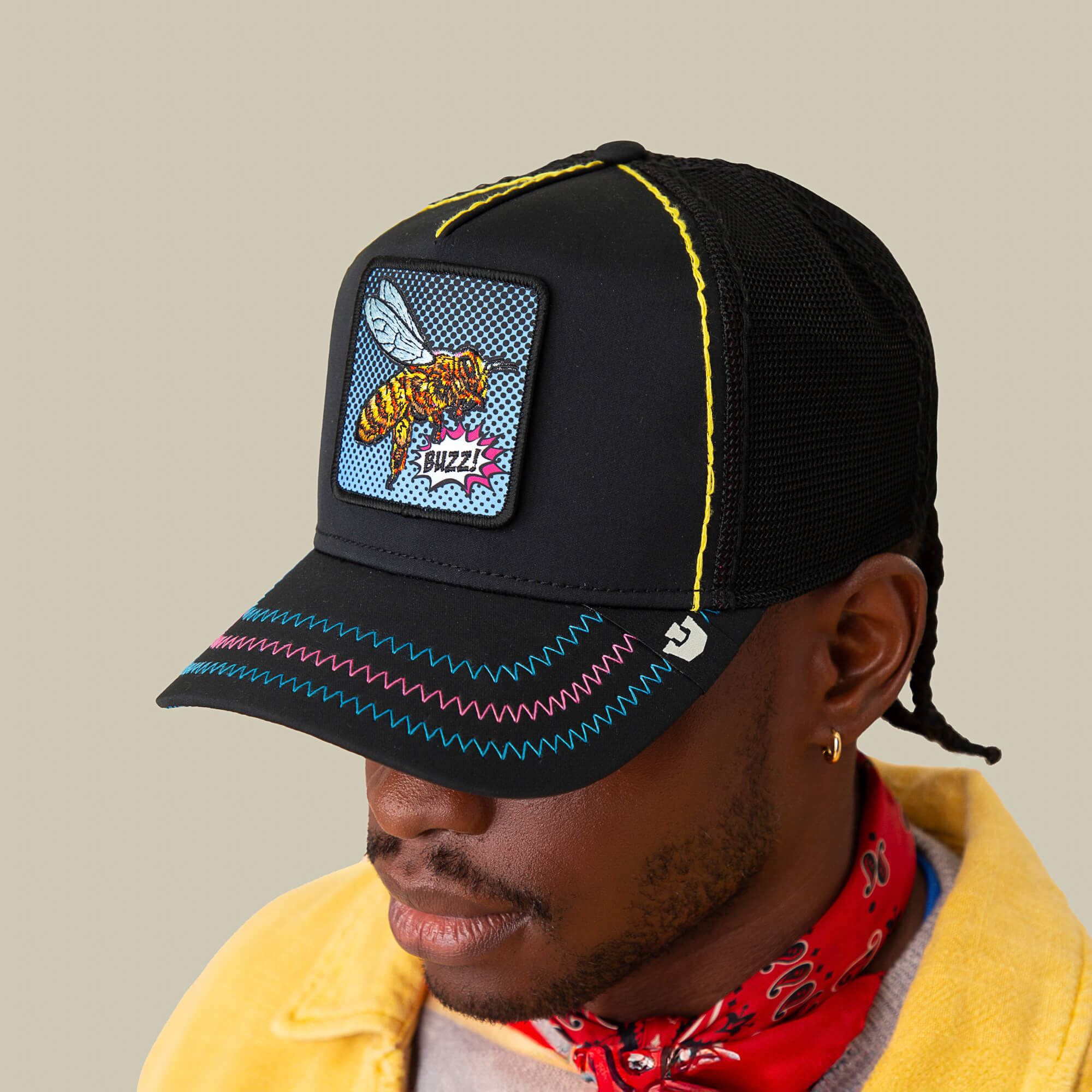 A person wears the Goorin Bros. "Oh Bee Hive" black mesh trucker hat featuring a pop-art bee patch and colorful stitching, styled with a yellow jacket and red bandana.