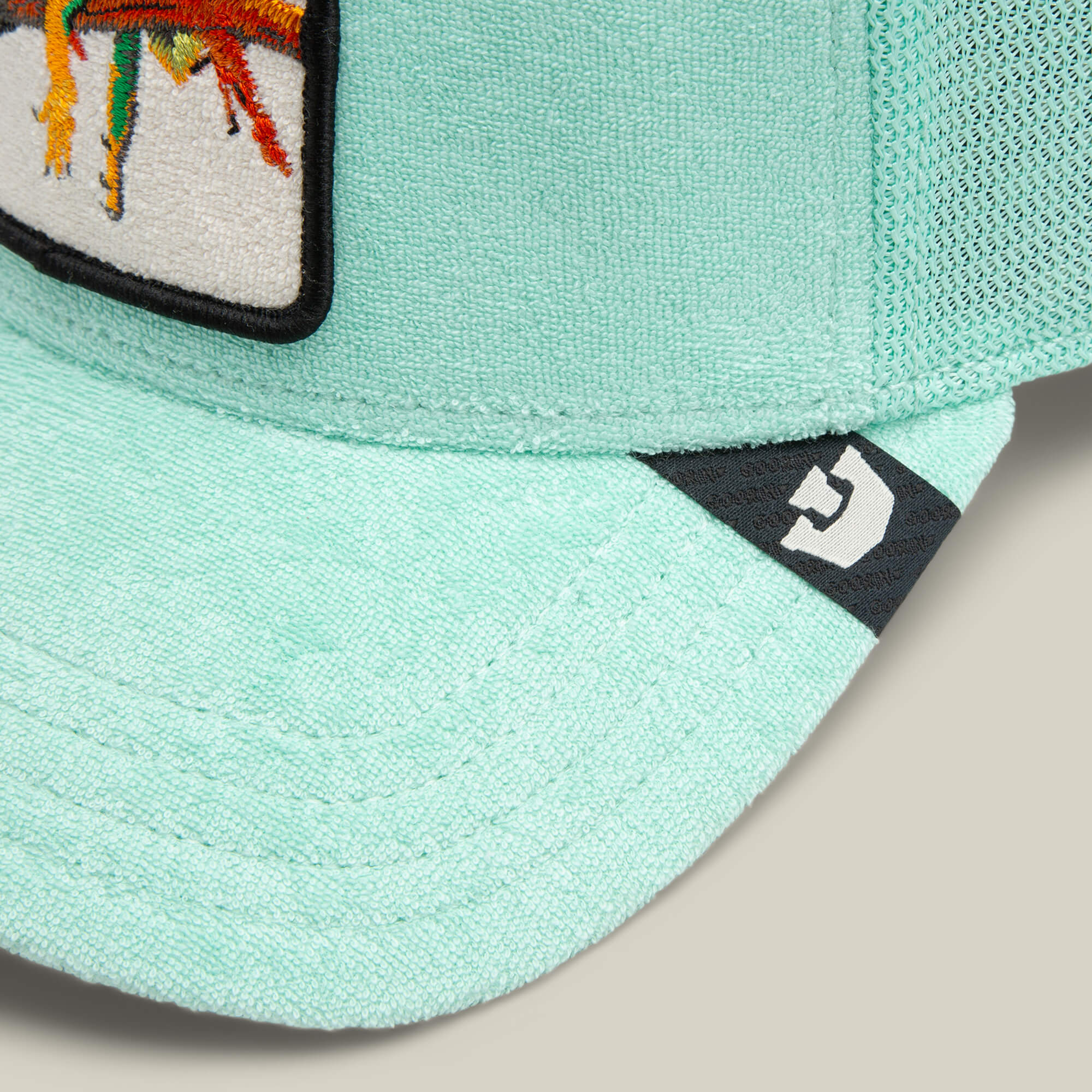 Close-up of the Terry Cloth Iguana cap by Goorin Bros.—a light teal, breathable hat with a mesh back, fabric patch featuring a colorful design, and a small white geometric logo on the brim.