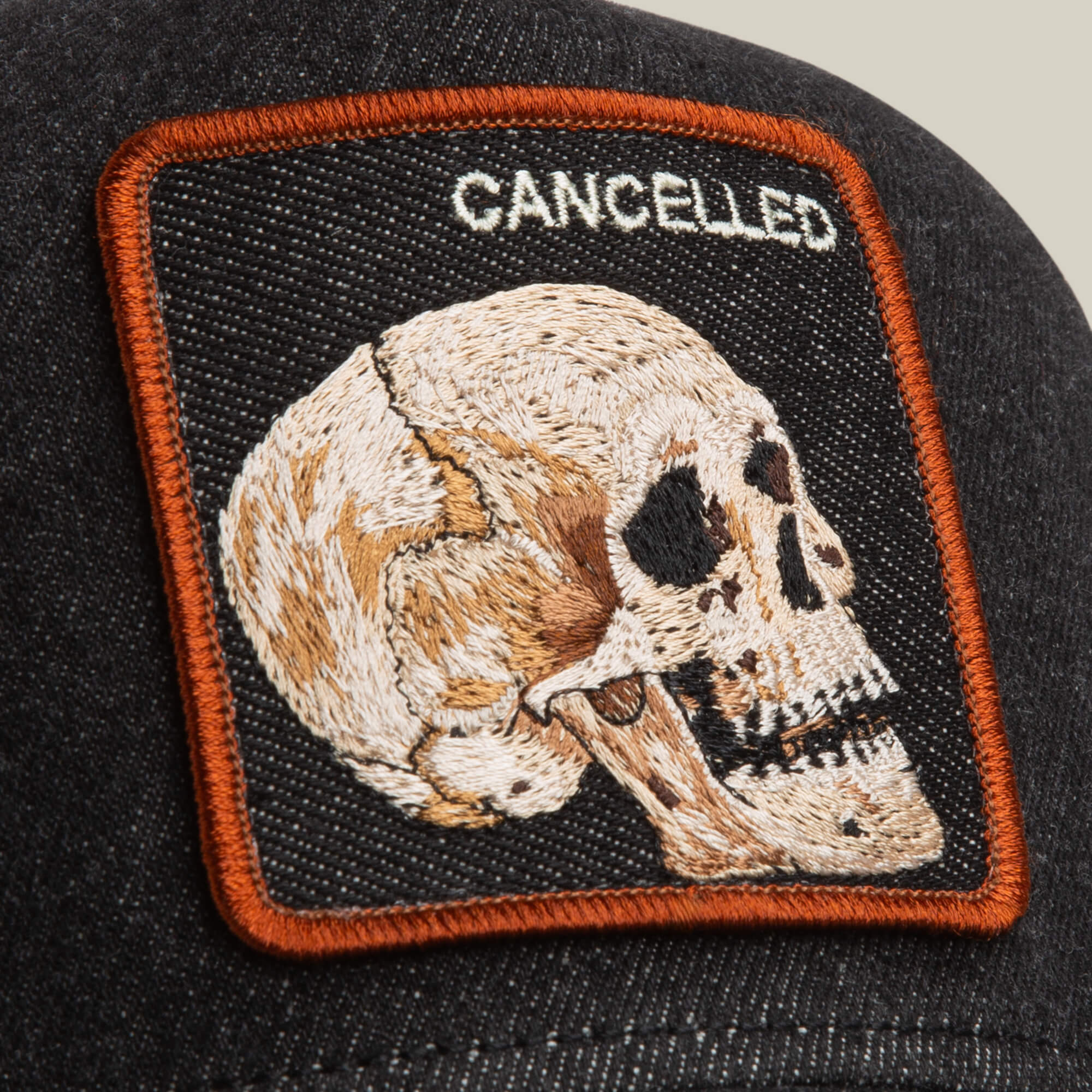 A close-up of the Goorin Bros. Denim Skull hat, a black denim cap with an embroidered skull patch and "CANCELLED" above it, outlined in orange thread.