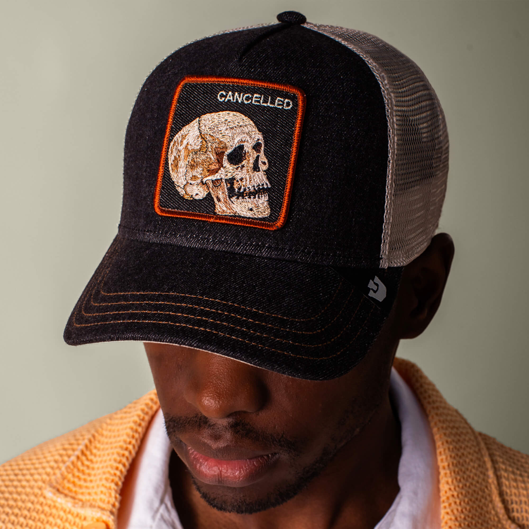 A man looks downward wearing the Goorin Bros. Denim Skull black and white mesh trucker cap, featuring a skull patch with “CANCELLED” embroidered above. He pairs it with a yellow textured shirt and dark wash black denim.