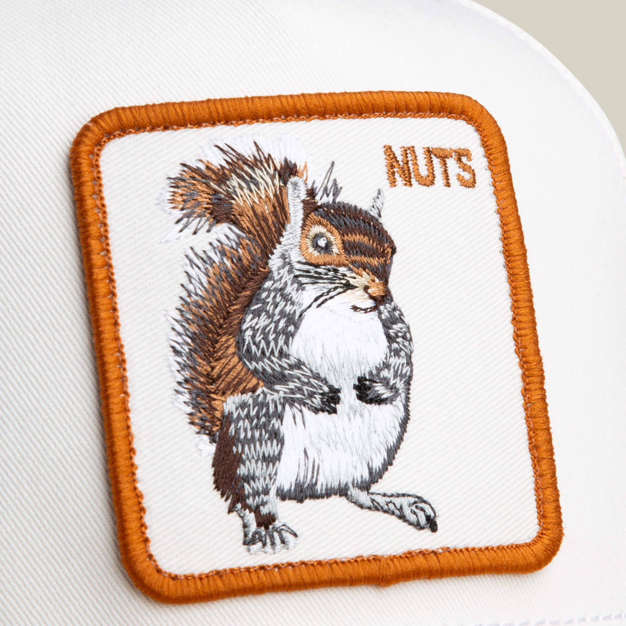 The Goorin Bros. Suede Squirrel cap features a white patch with an embroidered squirrel and bold orange "NUTS" lettering, accented by orange trim for playful charm on its white and tan design.
