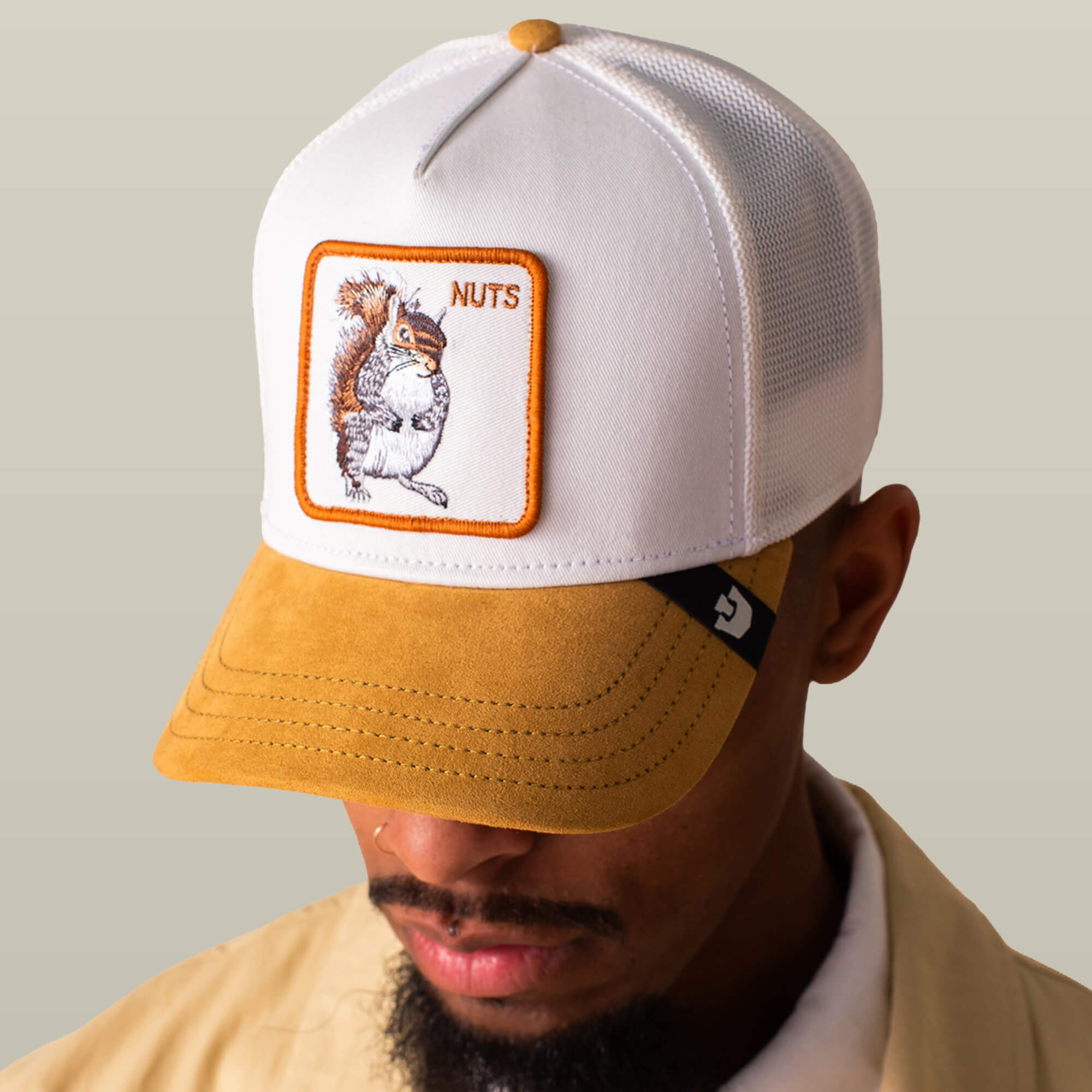 Man wears the Suede Squirrel hat by Goorin Bros., featuring a tan suede brim, white crown, and "NUTS" patch with a squirrel, set against a neutral background.