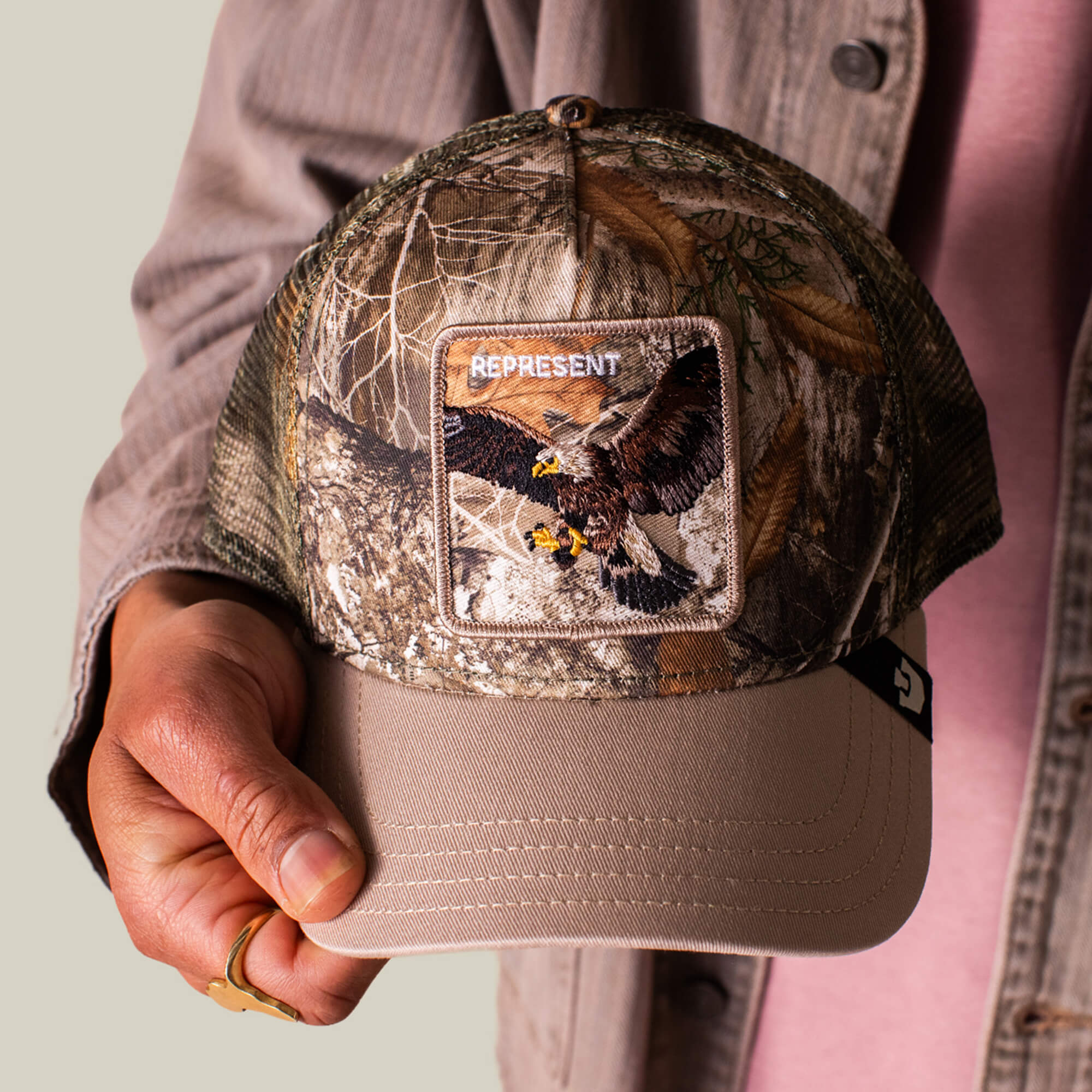 Someone in a beige jacket holds the Goorin Bros. Realtree Edge Represent Eagle trucker hat, featuring an embroidered eagle patch.