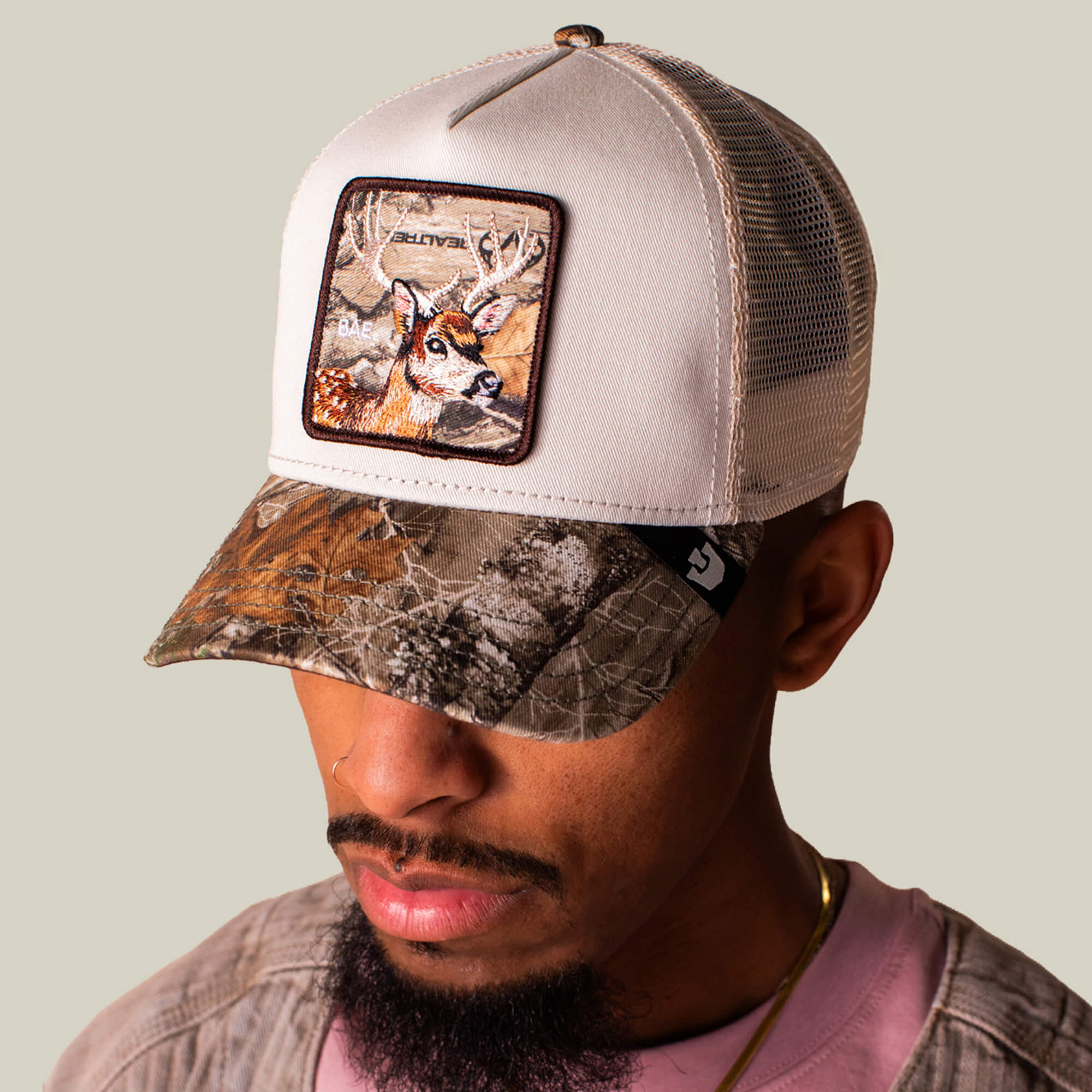 A man wears the Goorin Bros. Realtree Edge Bae Doe hat—a beige mesh cap with a Realtree Edge camo visor and a deer patch—while looking down, sporting a nose ring.