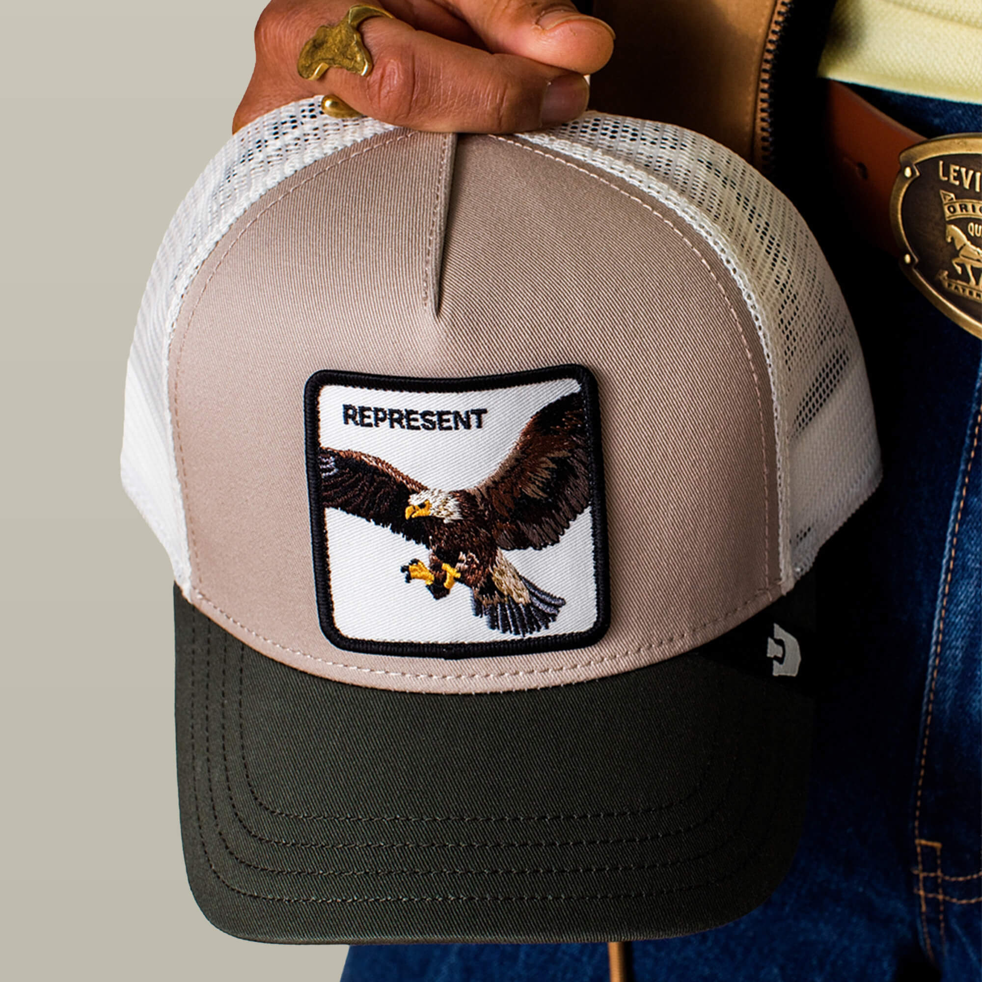 A person holds the Goorin Bros. Represent Eagle Trucker hat, which features an eagle patch and "REPRESENT" embroidery. They're also wearing a gold ring and a belt with a large buckle.