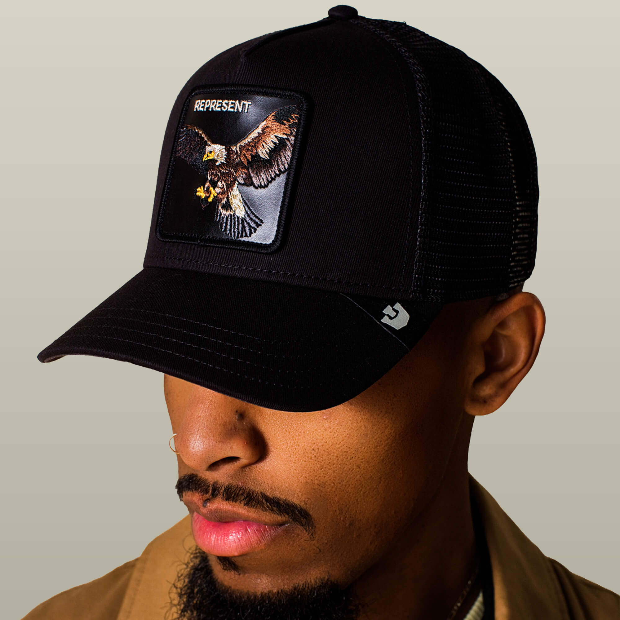 A man wears the Goorin Bros. Represent Eagle Trucker cap, which features an eagle patch with "REPRESENT" embroidered above it, set against a beige background.
