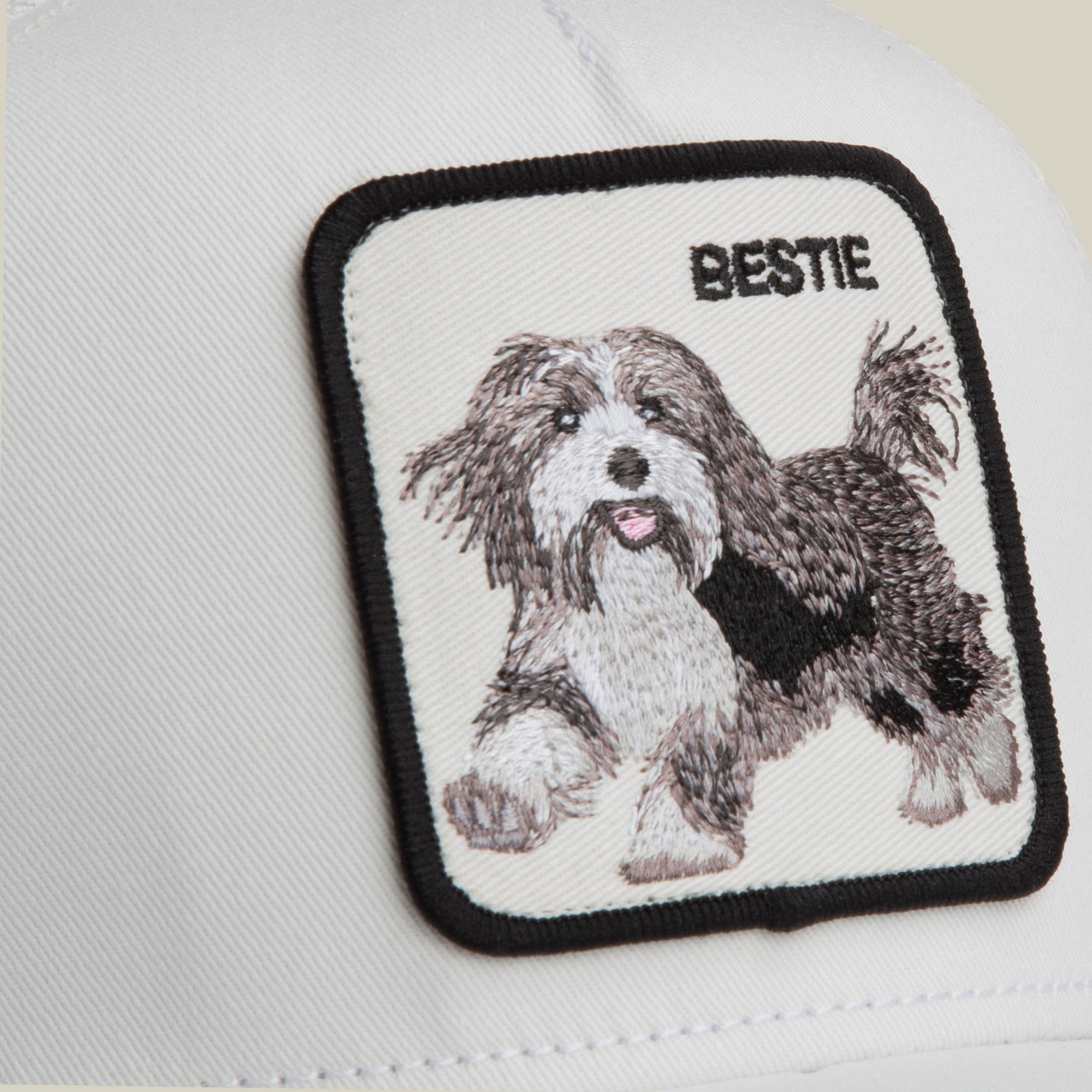 Close-up of the Goorin Bros. Bestie Trucker hat, white fabric with a square embroidered black and white dog patch labeled "BESTIE" in the top right corner.
