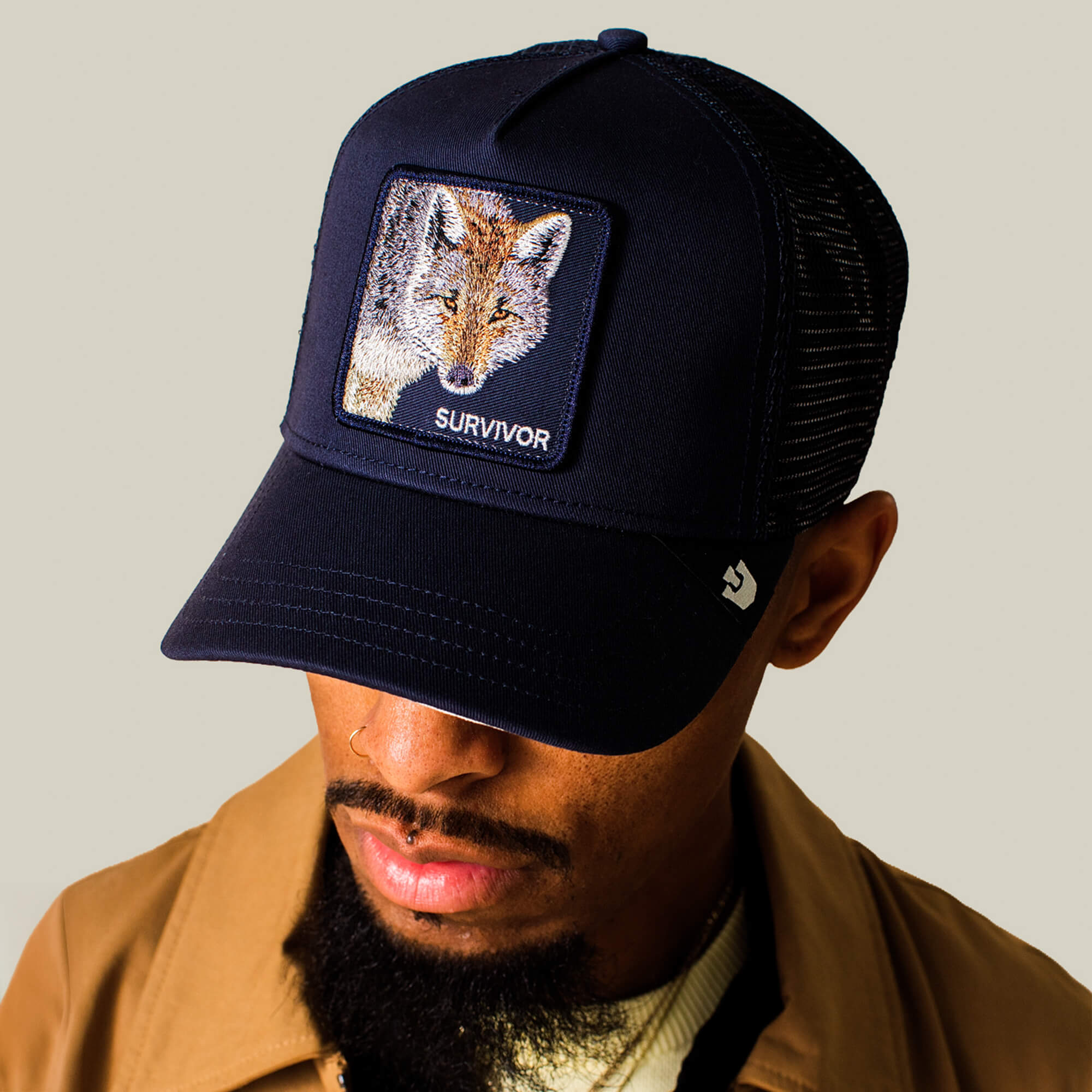 A man in a Goorin Bros. Mirage Survivor navy trucker hat with a fox patch, tan jacket, and white shirt looks downward against a plain background.