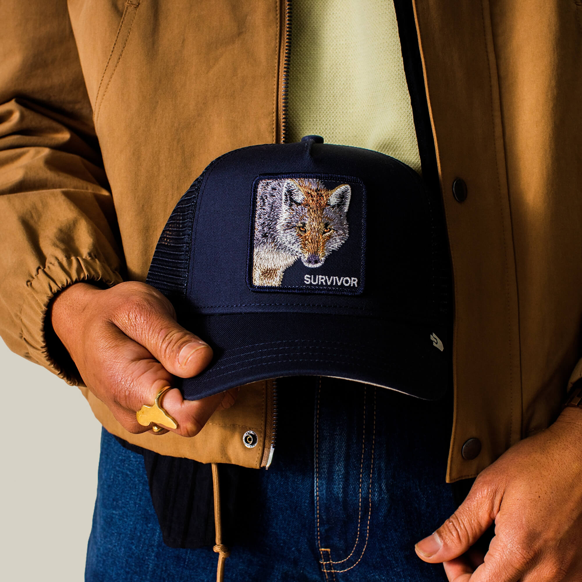A person in a brown jacket and blue jeans holds the Goorin Bros. Mirage Survivor cap, which features an embroidered fox patch and the word SURVIVOR.