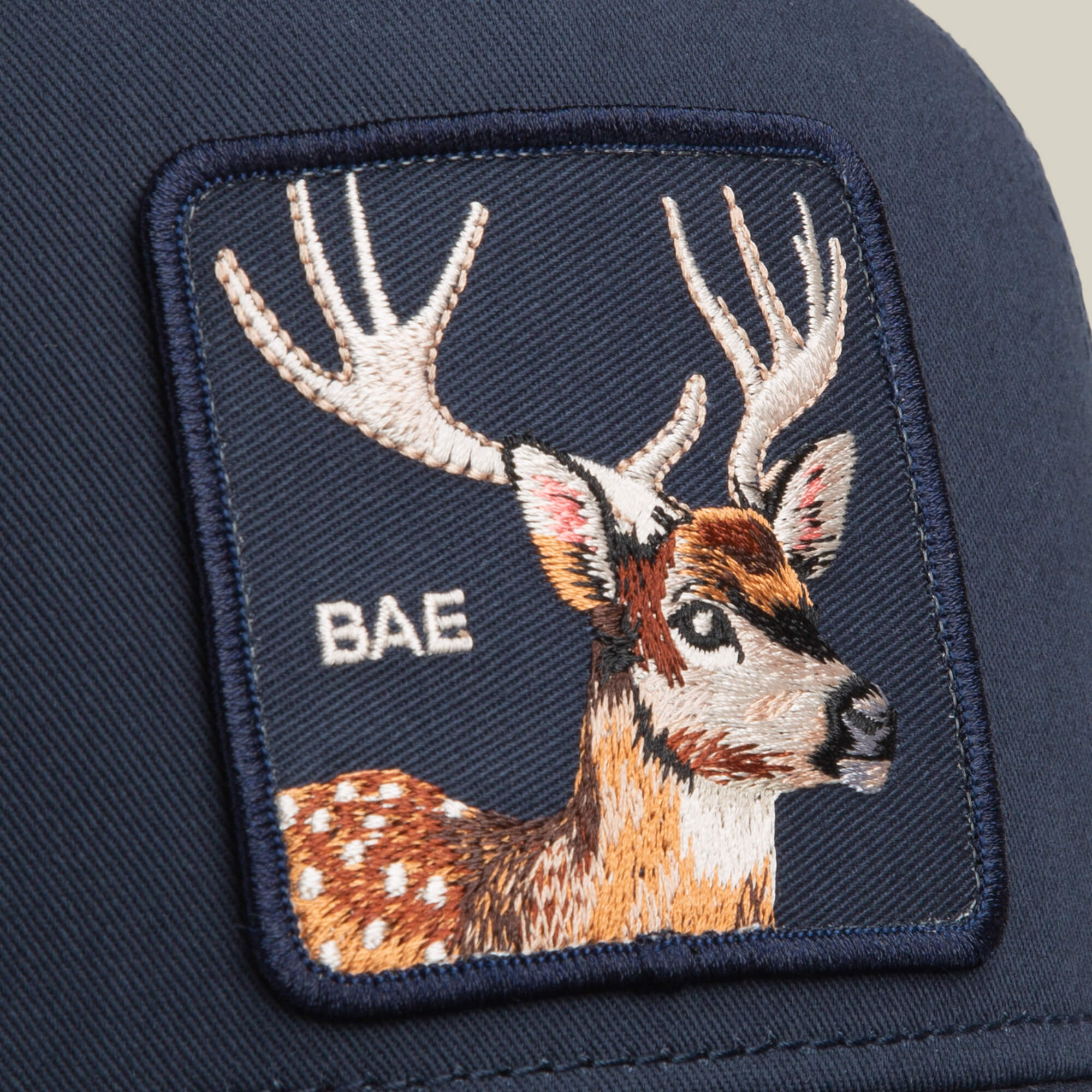 Close-up of the Goorin Bros. Mirage Bae navy trucker cap, showcasing an embroidered deer patch with antlers and "BAE" stitched on the front.