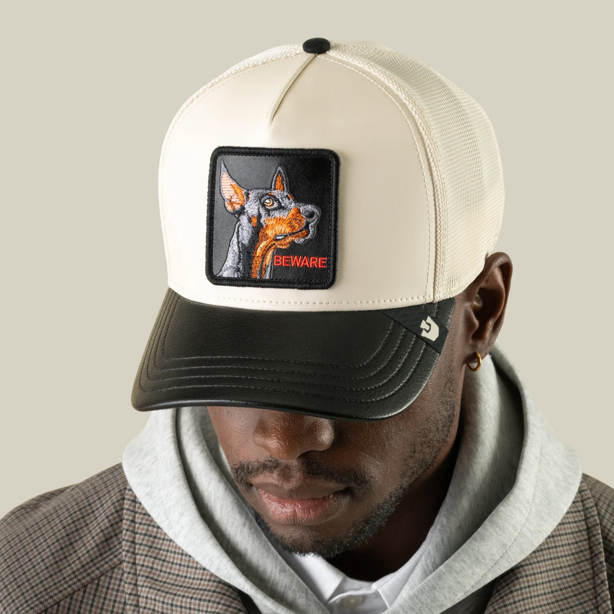 A man wearing the Goorin Bros. Faux Leather Doberman cream embroidered trucker hat with a BEWARE patch, paired with a gray hoodie and plaid coat, looks downward.