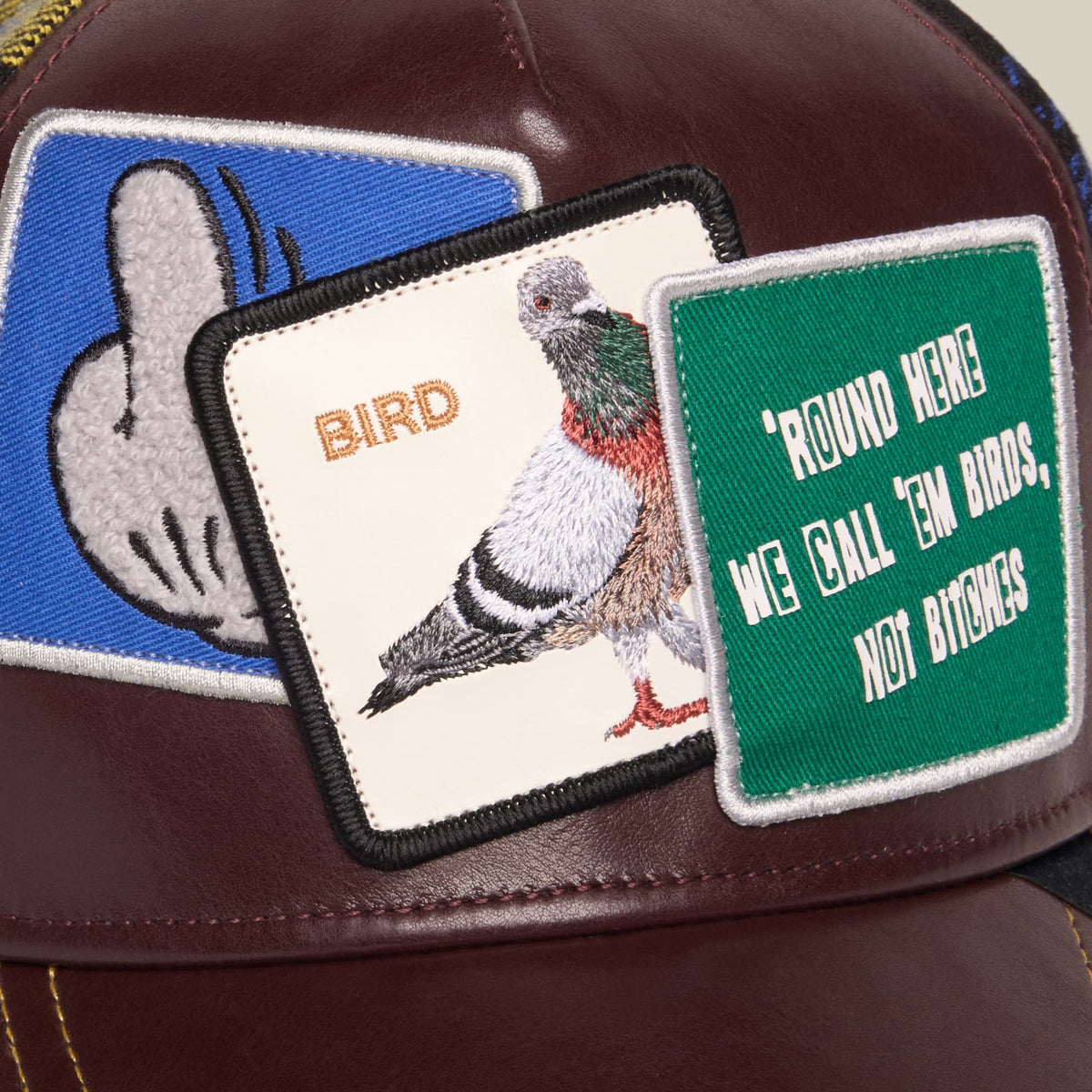 The Goorin Bros. Bird! trucker hat features a burgundy leather front panel, blue mesh side panel, and embroidered patches including a detailed bird illustration and green "Round Here We Call 'Em Birds, Not Bitches" text patch.