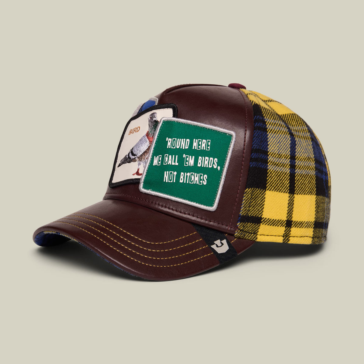The Goorin Bros. Bird! trucker hat features a burgundy leather front with a green patch reading "Round Here We Ball, Em Birds, Not Bitches," paired with a yellow and navy plaid fabric back, shown against a neutral studio background.