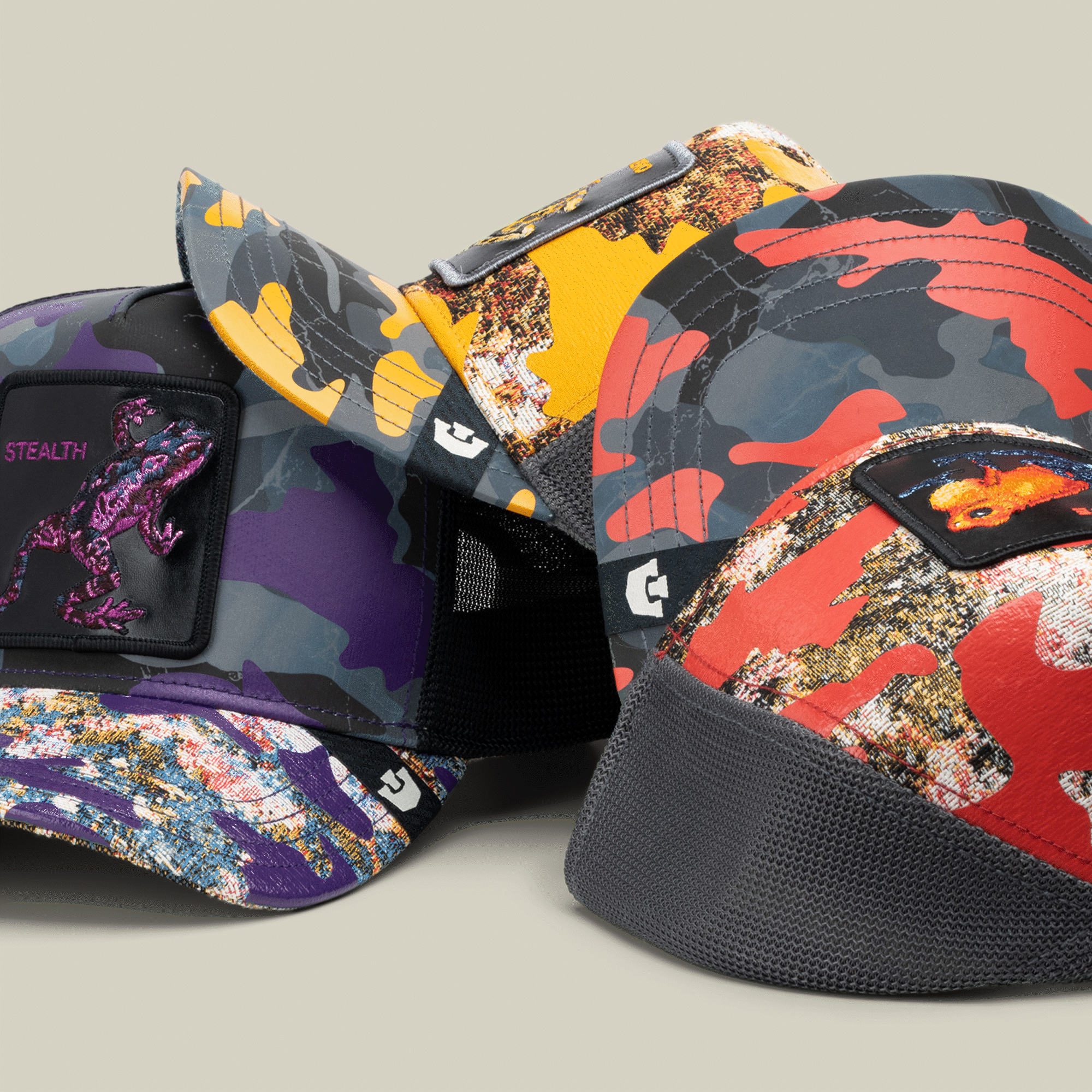 Four colorful camo baseball caps by Goorin Bros. are shown, including the standout Silent But Deadly purple trucker hat—each with a unique embroidered dinosaur patch on the front panel and stylish mesh backs.