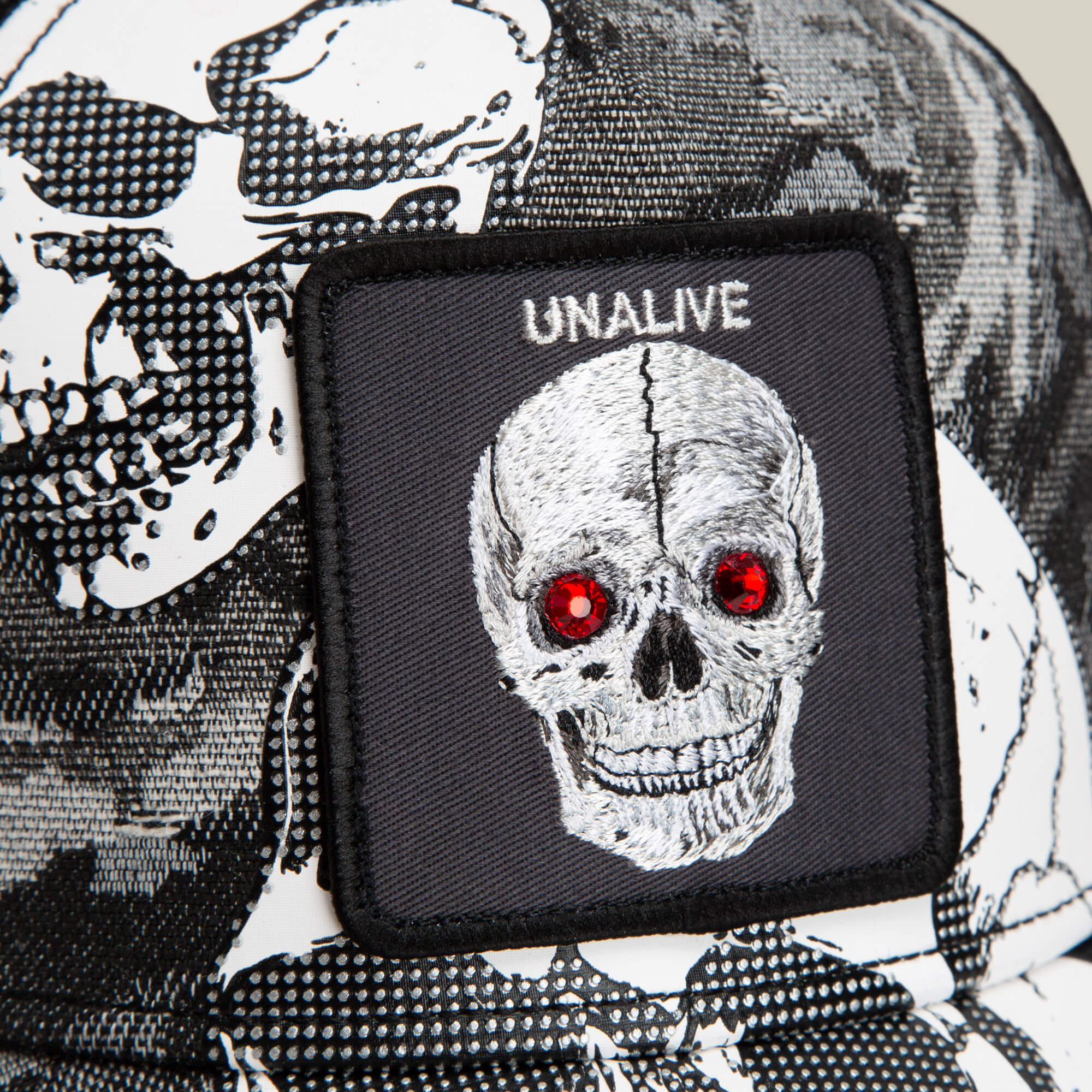 Close-up of the Goorin Bros. Bone Head Unalive trucker hat, showcasing a patch with an embroidered skull featuring red eyes and the word “UNALIVE,” a black-and-white skull pattern on the fabric, and a breathable mesh back for added comfort.