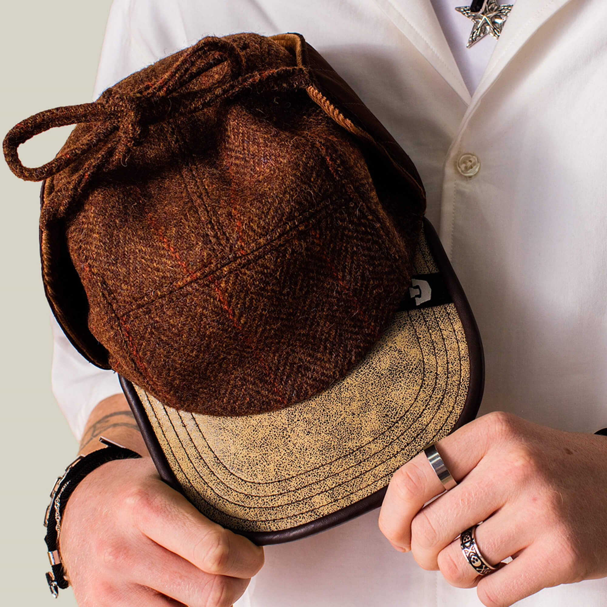 A person in a white shirt holds the Goorin Bros. Frazier Lake brown wool trapper cap with ear flaps and a beige brim, displaying rings and bracelets on their hands.