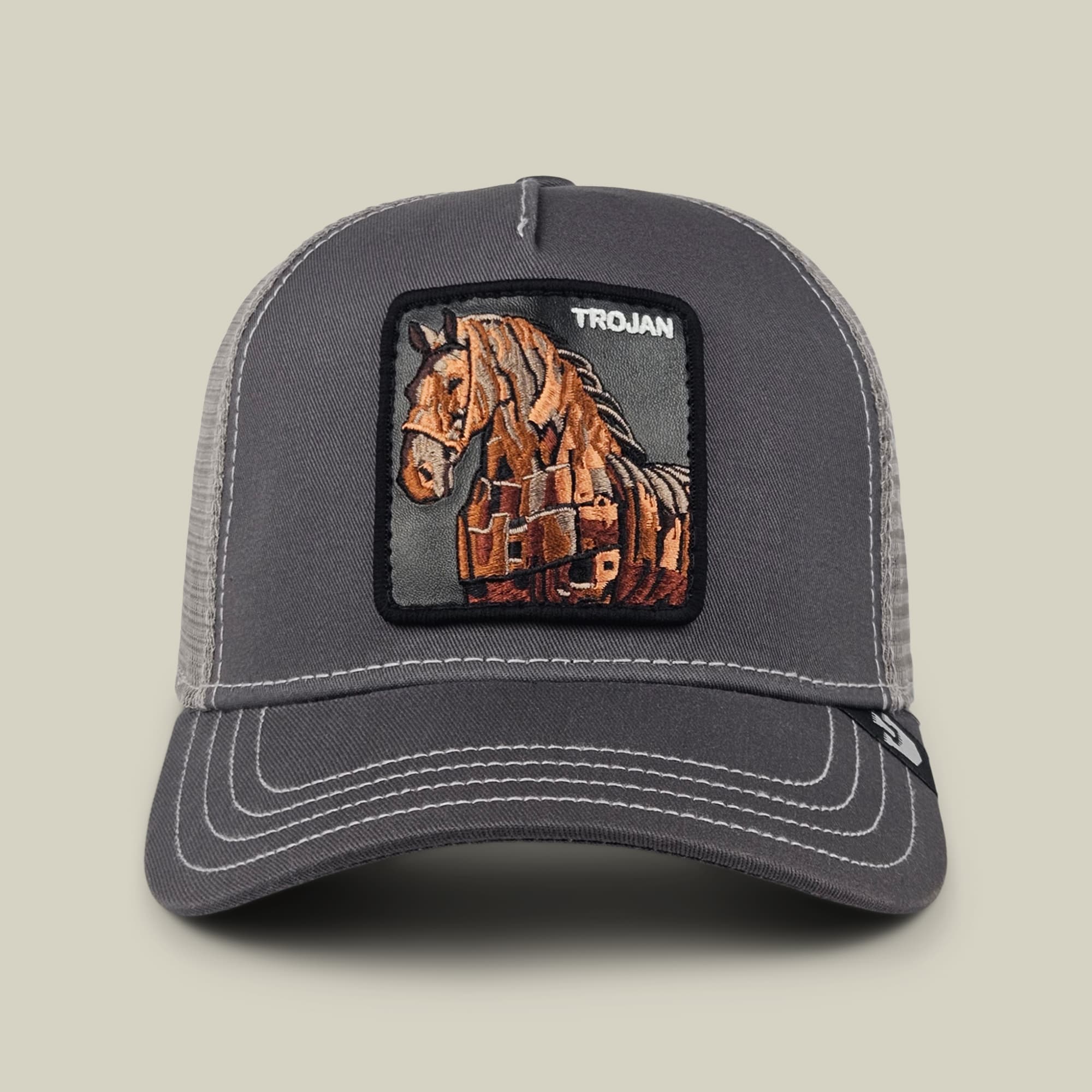 The Goorin Bros. 99 Strong trucker hat features a charcoal gray mesh back, dark front panel, and embroidered horse patch in brown and orange with text reading "Trojan" displayed on a neutral studio background.