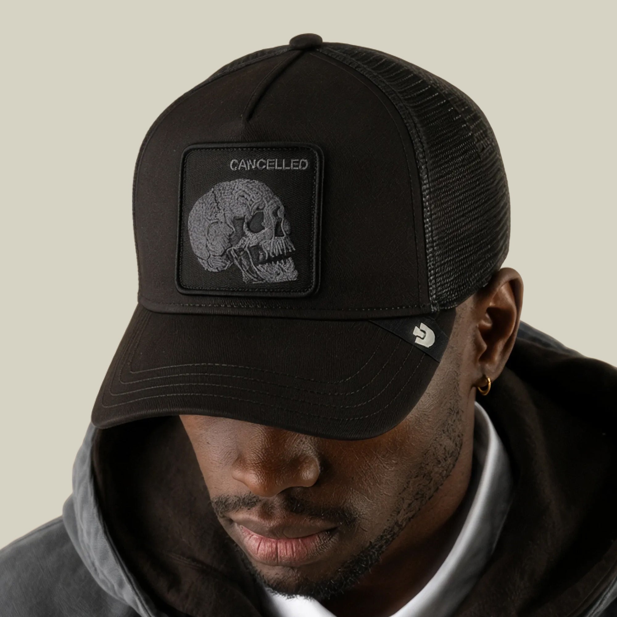 A man wearing the Goorin Bros. Tonal Skull trucker hat, featuring a matte dark finish and a skull patch with the word “CANCELLED,” looks down while dressed in a hooded jacket.