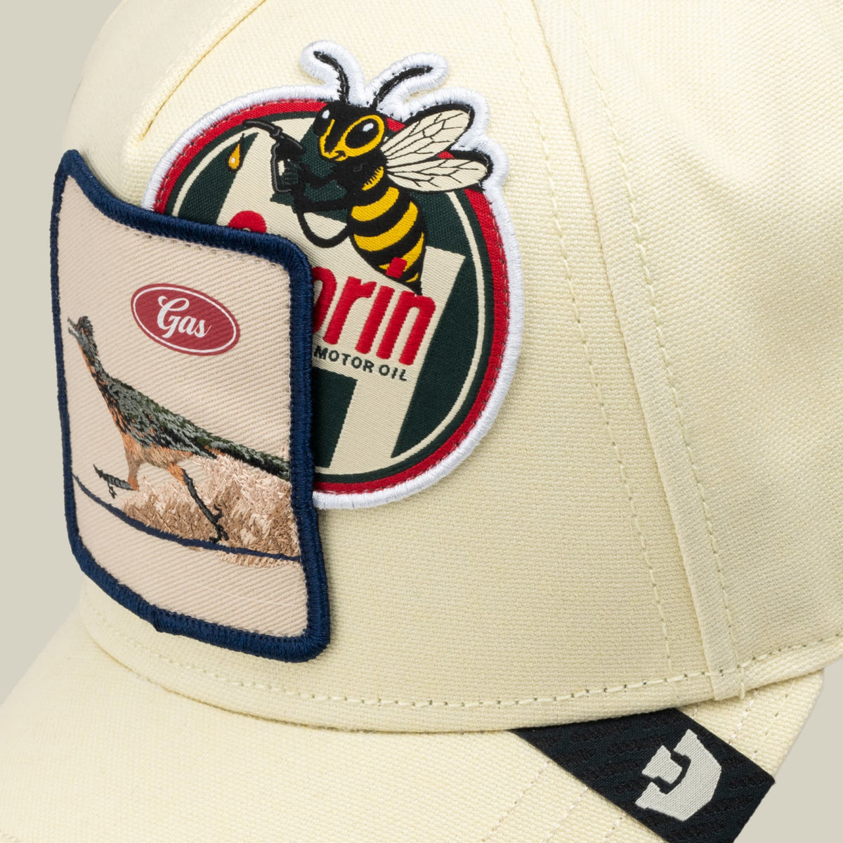 The Crude Guzzler by Goorin Bros is a cream-colored trucker cap with navy trim, featuring embroidered vintage-style patches including a bee logo and motor oil imagery on the front panel, displayed on a neutral background.