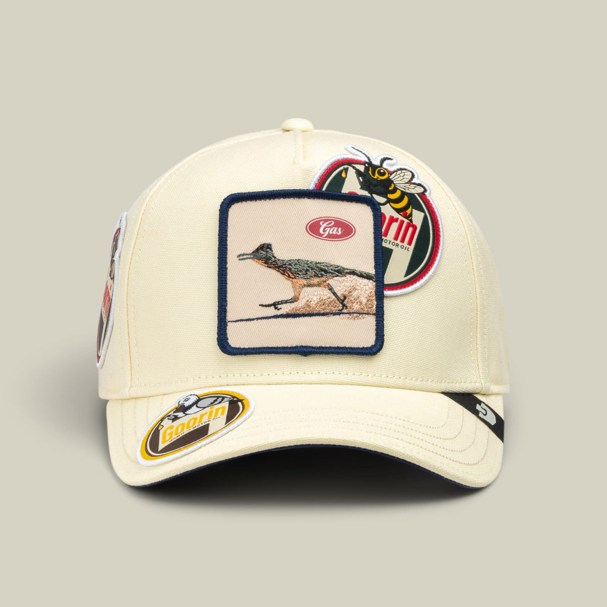 The Goorin Bros. Crude Guzzler is a cream-colored baseball cap featuring embroidered patches of a bird and a bee on the front and side, with navy trim and a curved bill against a neutral studio background.
