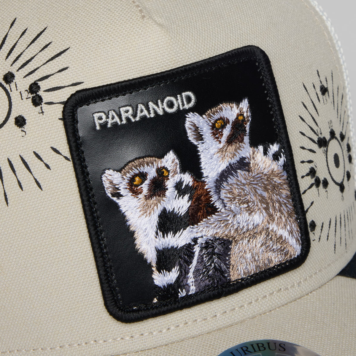 The Goorin Bros. G-Anon trucker hat features a cream canvas crown with black mesh back and an embroidered paranoid patch depicting two wide-eyed lemurs with golden eyes on a black background.