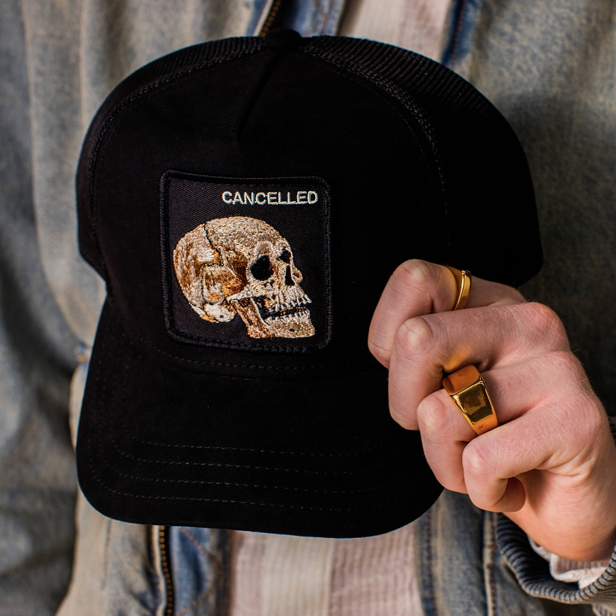 Someone is holding the Bold Suede Skull by Goorin Bros., a limited edition black microsuede trucker hat featuring a stitched skull patch with "CANCELLED" embroidered above it.