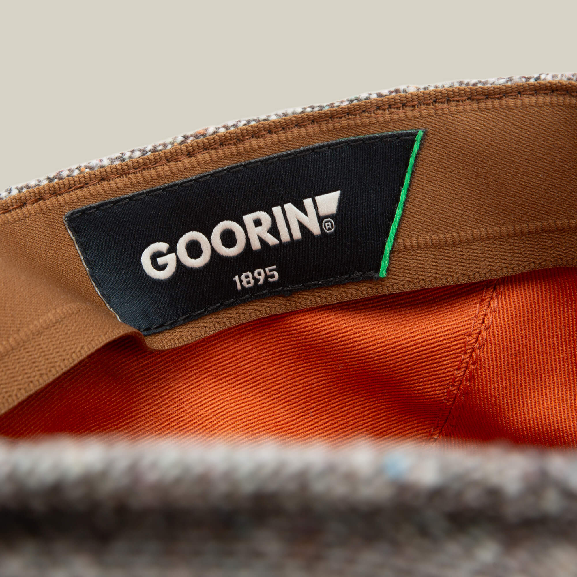 Close-up of the inside of a grey tweed Goorin Bros. Gibbins flatcap, featuring a black GOORIN 1895 label stitched onto brown and orange fabric—a classic detail in men's fashion.